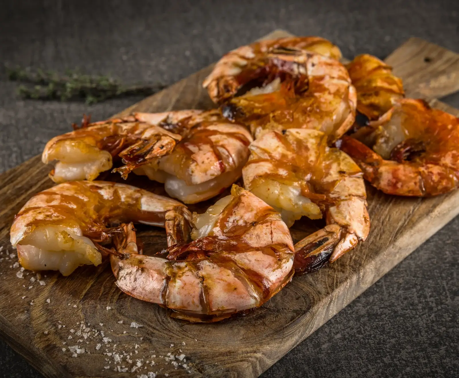 Nahaufnahme von mehreren goldbraun gegrillten Garnelen mit Schale auf einem rustikalen Holzbrett, bestreut mit grobem Meersalz. Im Hintergrund sind Kräuter und eine dunkle Oberfläche zu sehen.