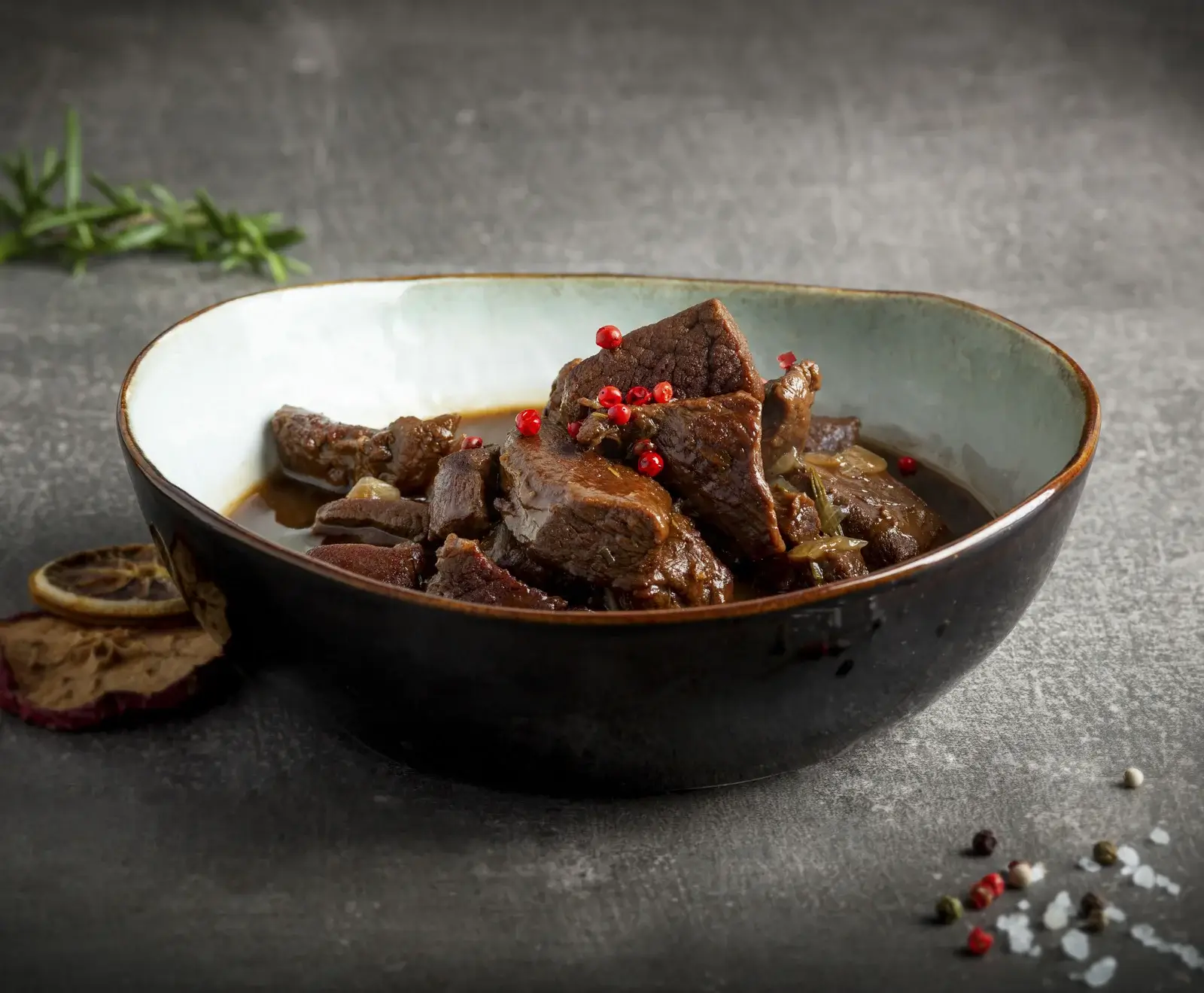 Eine Schale mit dunklem, geschmortem Fleisch (Gulasch oder Schmorbraten) in einer reichhaltigen Sauce, garniert mit rosa Pfefferbeeren. Daneben liegen getrocknete Zitronen- und Apfelscheiben, sowie Pfefferkörner und Salz.