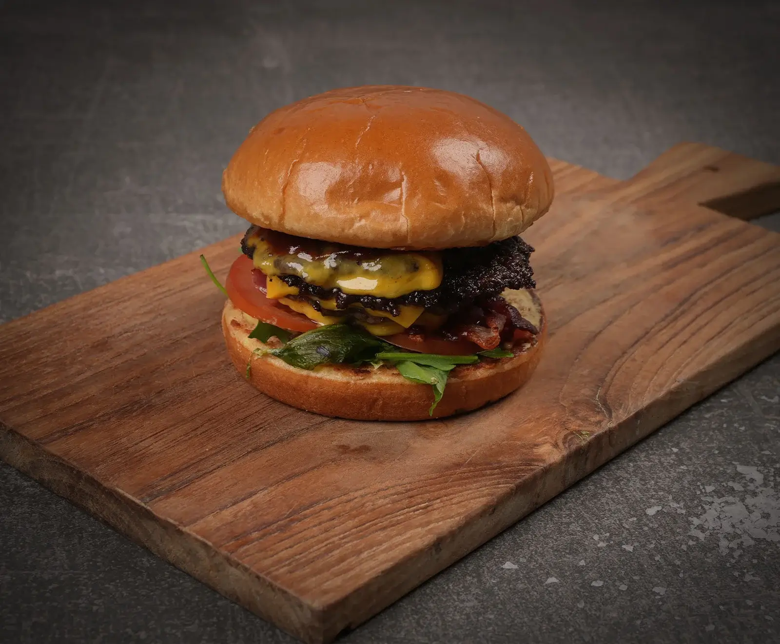 Ein saftiger Doppel-Cheeseburger mit glänzendem Brioche-Brötchen, zwei Rindfleisch-Patties, geschmolzenem Käse, knusprigem Speck, frischen Tomaten und grünem Salat, serviert auf einem Holzbrett.