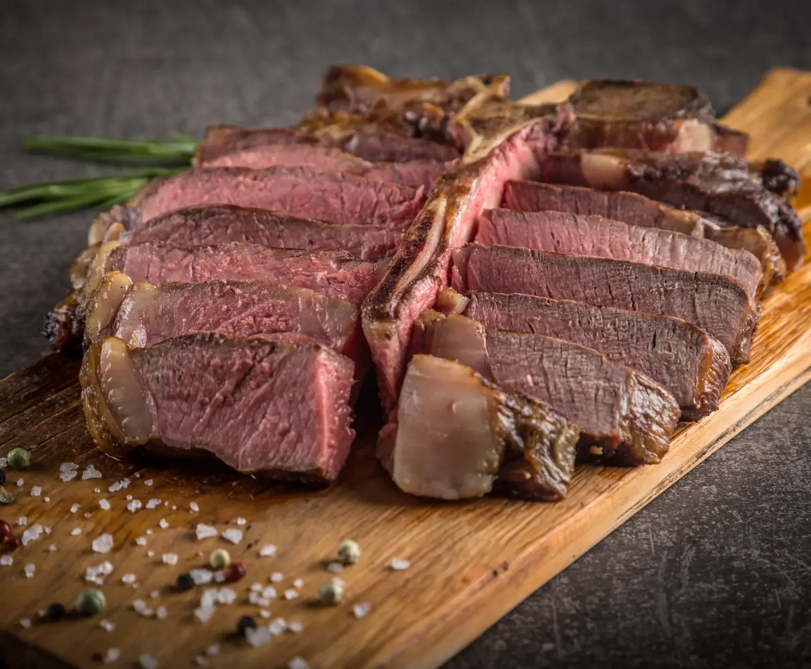 Ein perfekt gegartes, geschnittenes T-Bone-Steak auf einem Holzschneidebrett. Das Fleisch ist medium-rare mit rosa Kern, daneben liegen grobes Salz, bunte Pfefferkörner und Rosmarin.