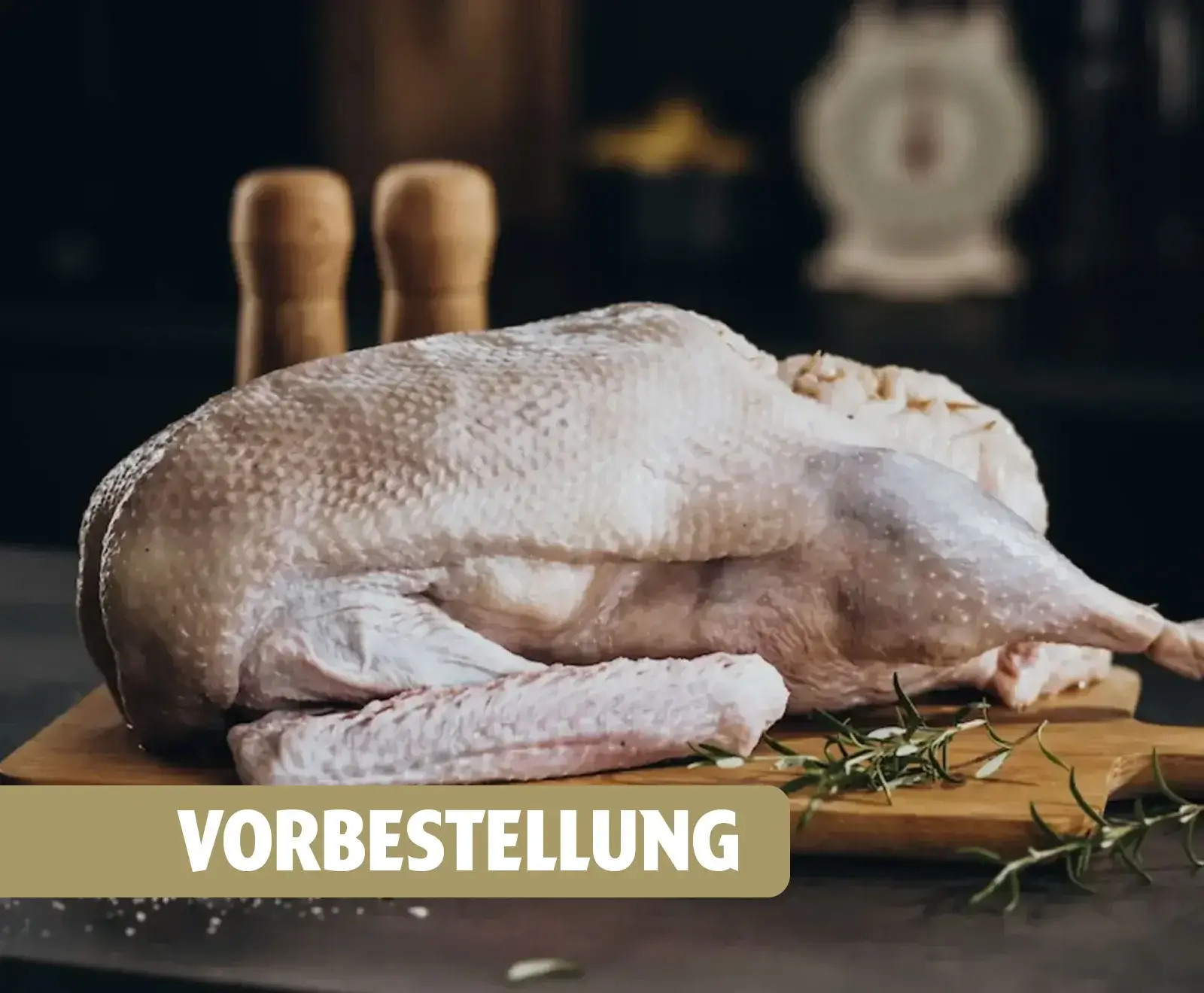 Eine rohe, gerupfte Ente oder Gans liegt auf einem Holzbrett mit Rosmarin. Im unscharfen Hintergrund sind Gewürzmühlen und eine Küchenwaage zu erkennen. Ein Textbanner mit 'VORBESTELLUNG' ist eingeblendet.