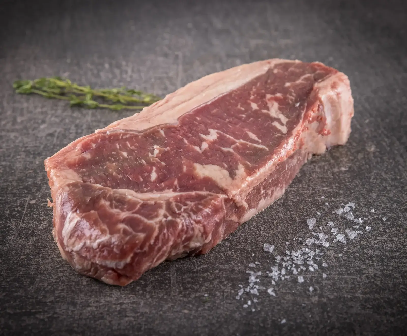 Nahaufnahme eines rohen, marmorierten Rindersteaks mit Fettrand auf einer dunklen Oberfläche, begleitet von Thymianzweigen und grobem Meersalz.