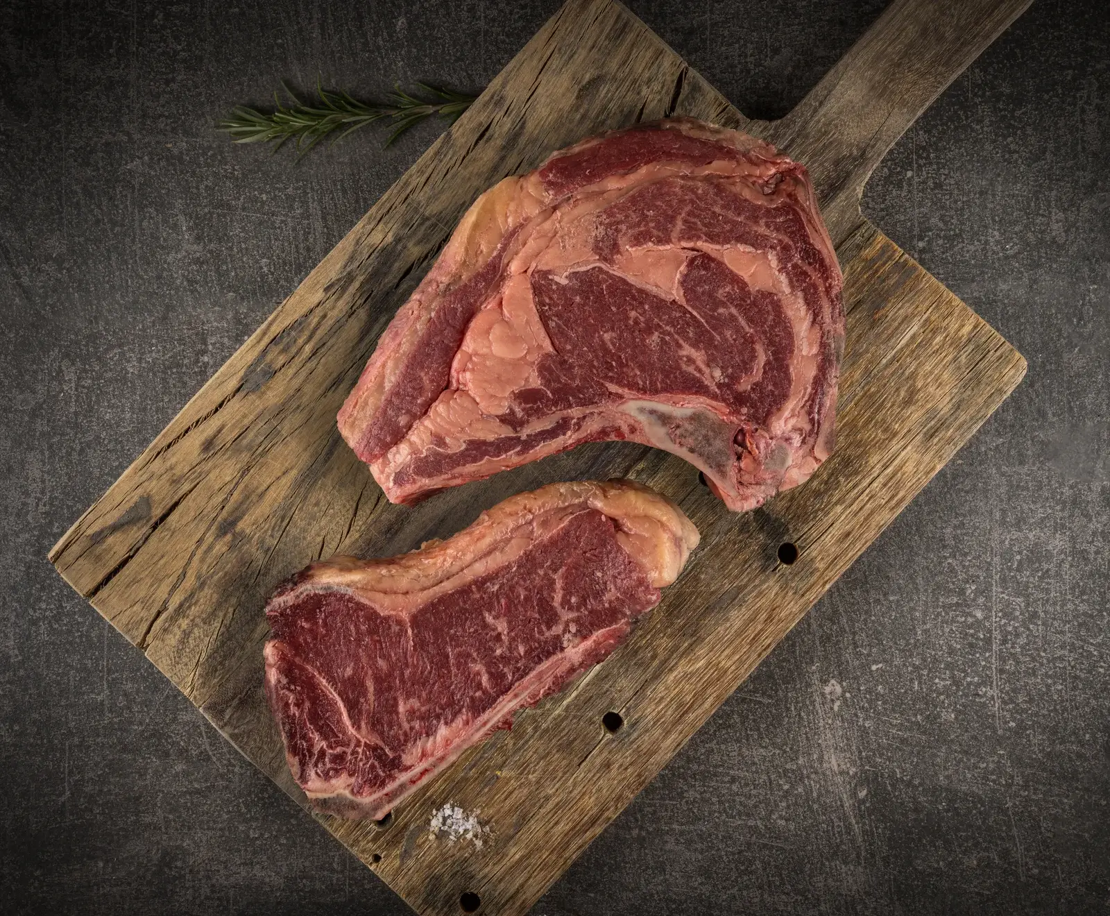 Zwei rohe Rindersteaks (ein Ribeye mit Knochen, ein kleineres ohne) auf einem rustikalen Holzbrett, mit Rosmarin und grobem Salz auf dunklem Hintergrund.