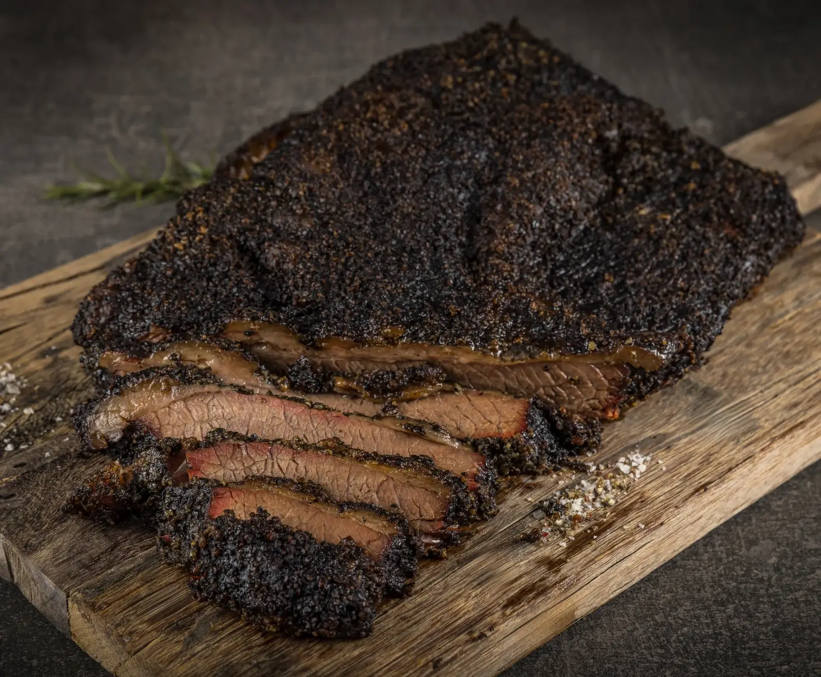 Ein großes, aufgeschnittenes Stück geräuchertes Rinderbruststück (Brisket) mit dunkler Kruste und rosa Rauchring, angerichtet auf einem rustikalen Holzbrett mit Salz, Pfeffer und einem Rosmarinzweig.