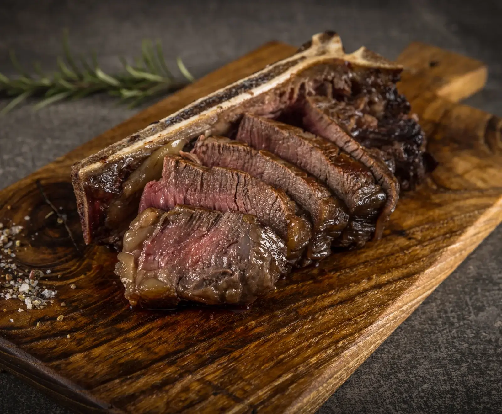 Ein geschnittenes T-Bone-Steak, medium-rare, auf einem rustikalen Holzbrett. Das saftige Fleisch zeigt eine rosa Mitte. Grobes Salz, Pfeffer und ein Rosmarinzweig sind sichtbar.