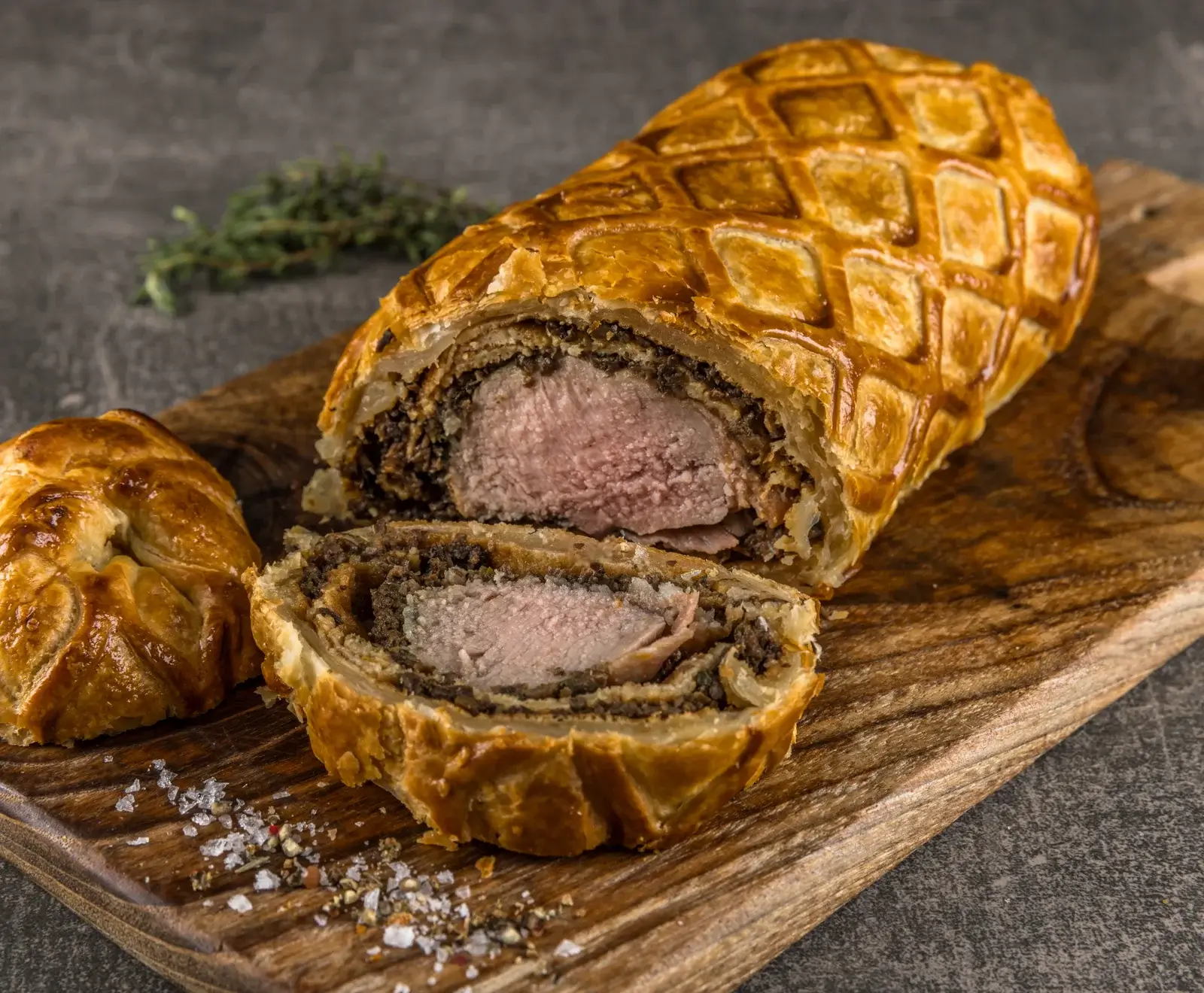 Ein angeschnittenes Beef Wellington auf einem Holzbrett. Der goldbraune Blätterteig mit Rautenmuster umschließt rosa Rinderfilet und dunkle Pilz-Duxelles. Grobes Salz und Pfeffer sind verstreut.