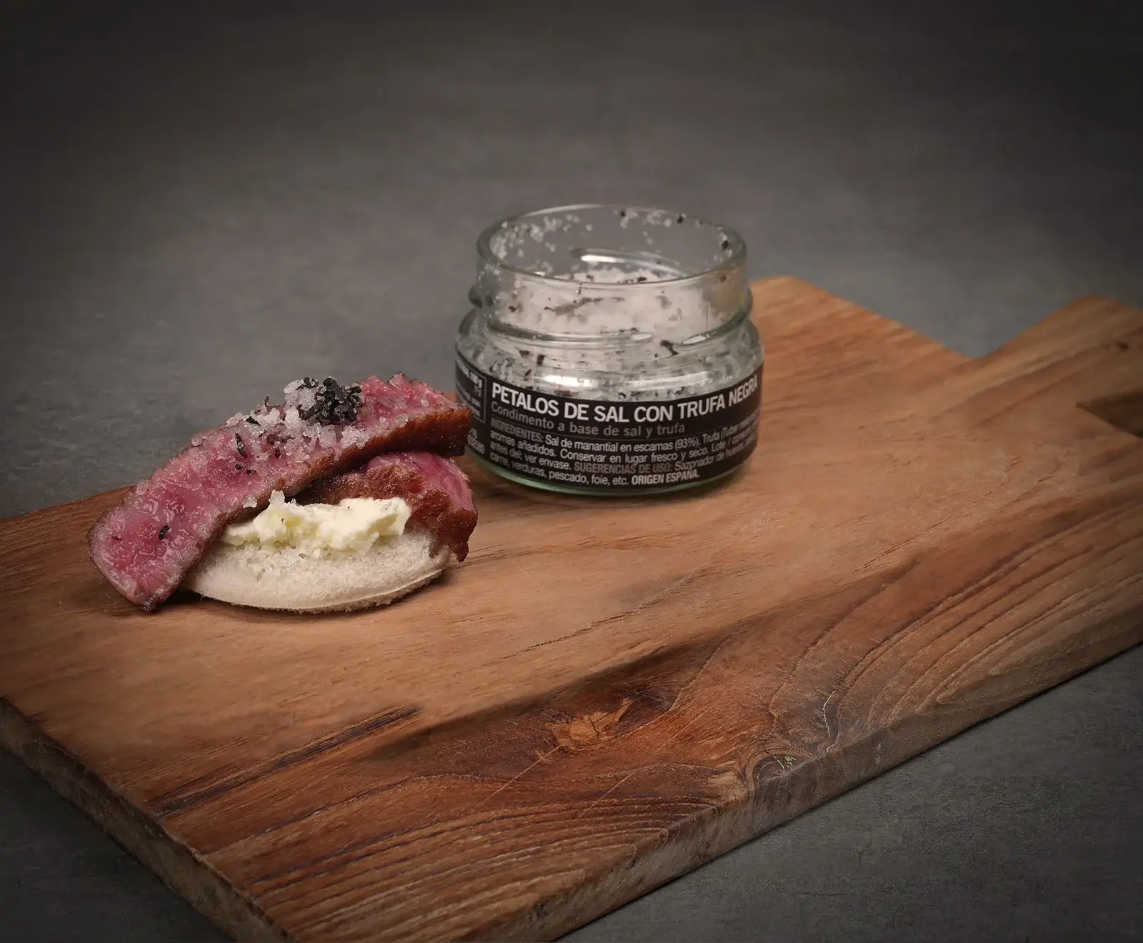 Ein appetitliches Häppchen mit rosa gebratenem Fleisch, Frischkäse und Trüffelsalz auf Brot, daneben ein Glas "Petalos de Sal con Trufa Negra" auf einem Holzbrett.