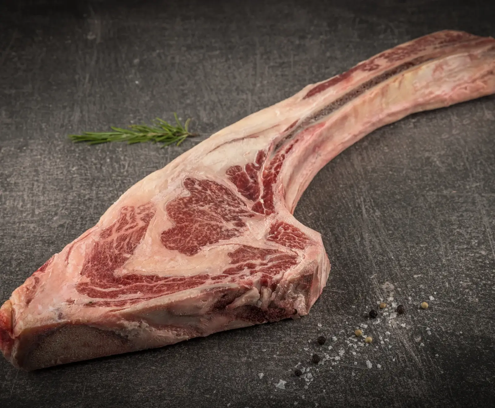 Ein rohes Tomahawk-Steak mit langem Knochen und ausgeprägter Marmorierung liegt auf einer dunklen, strukturierten Oberfläche. Daneben sind ein Rosmarinzweig, grobes Salz und Pfefferkörner zu sehen.