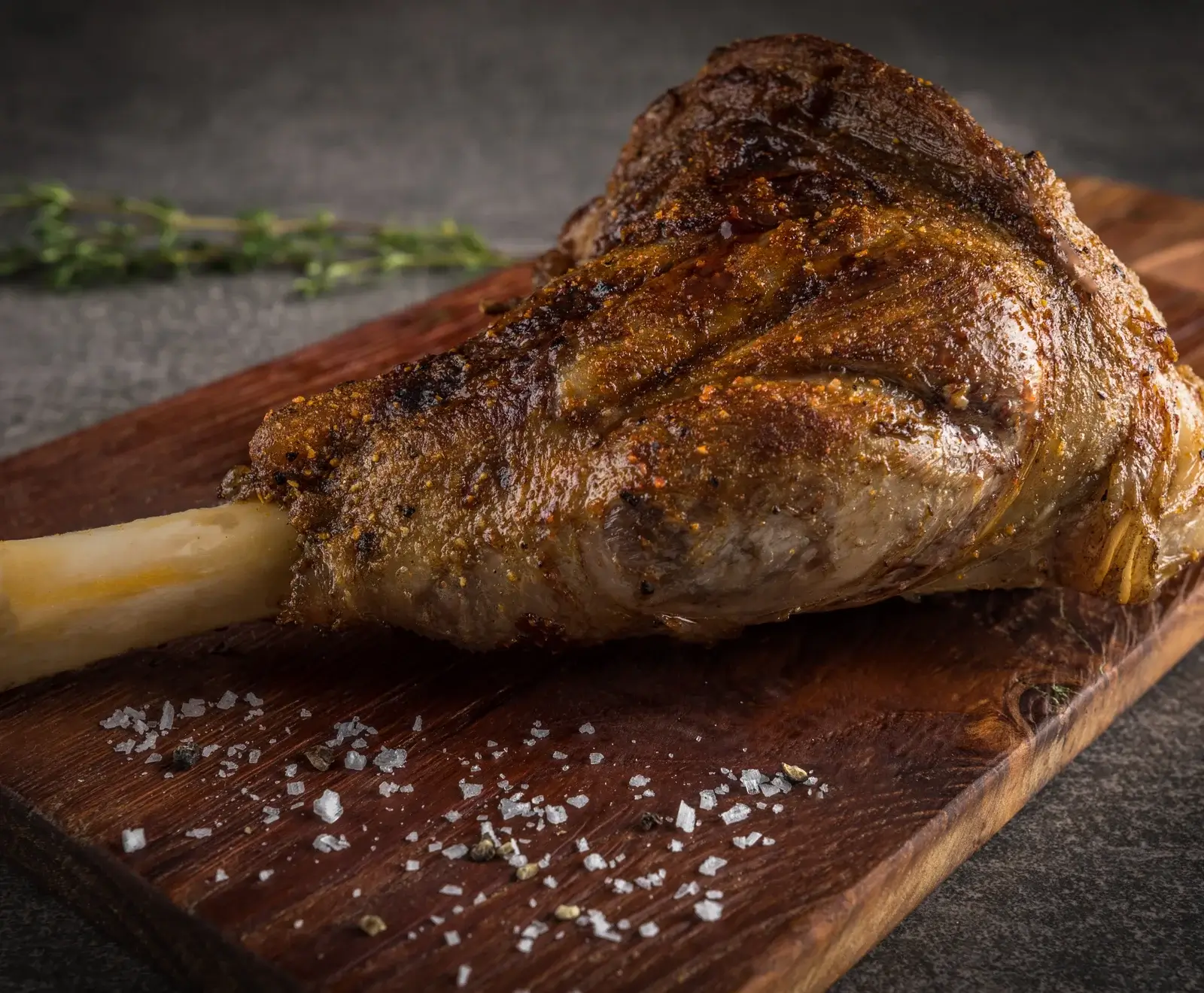 Ein knusprig gebratenes Lammhaxenstück mit goldbrauner Haut und Gewürzen, auf einem dunklen Holzbrett liegend. Grobe Salzkristalle und Pfeffer sind auf dem Brett verstreut, im Hintergrund frische Kräuter.