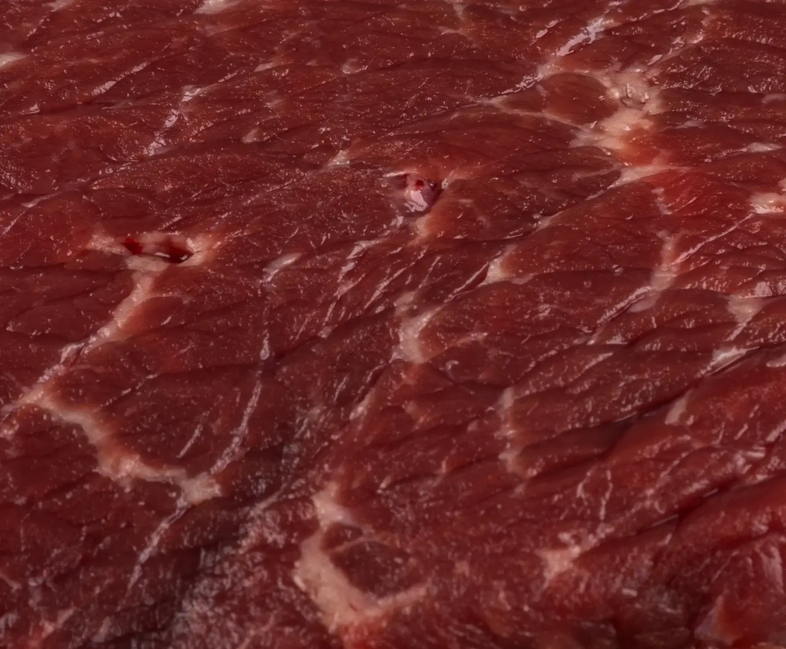 Nahaufnahme von frischem, rotem Rindfleisch mit ausgeprägter weißer Marmorierung durch Fettadern und sichtbaren Muskelfasern. Eine kleine, helle Stelle ist im Fleischgewebe erkennbar.