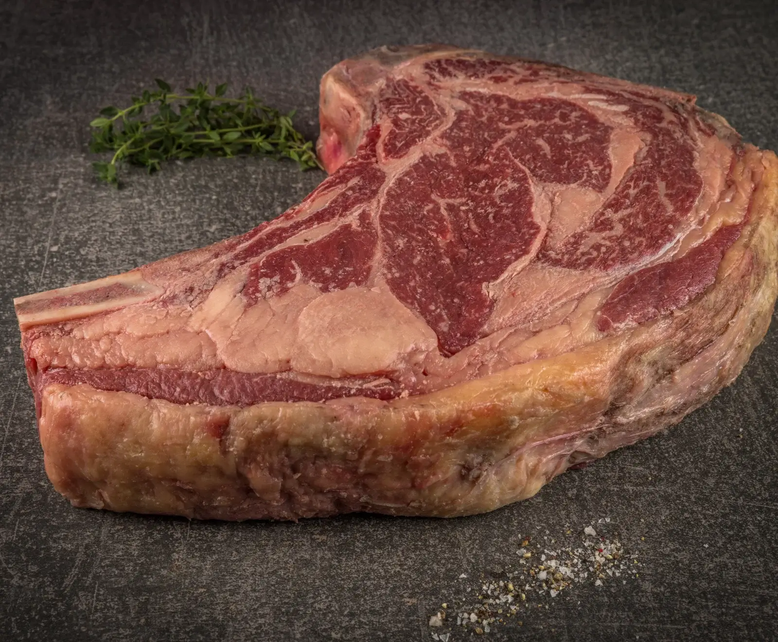 Ein großes, rohes Ribeye-Steak mit Knochen (Tomahawk) liegt auf einer dunklen, strukturierten Oberfläche. Das stark marmorierte Fleisch hat eine dicke Fettschicht. Links liegt frischer Thymian, rechts eine Prise Salz und Pfeffer.