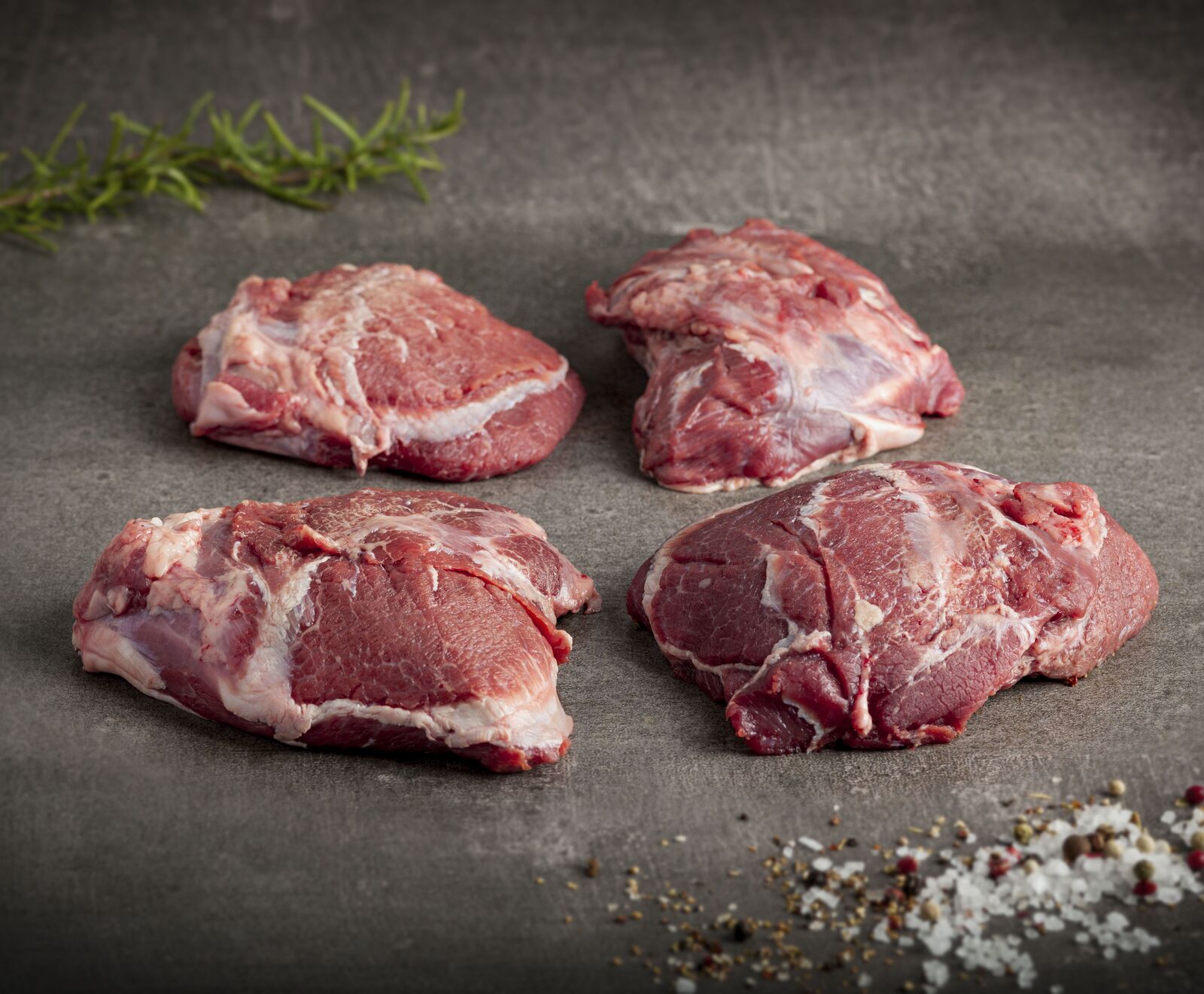 Vier rohe Lammkoteletts mit Rosmarin auf dunklem Hintergrund mit Salz und Pfeffer bestreut.