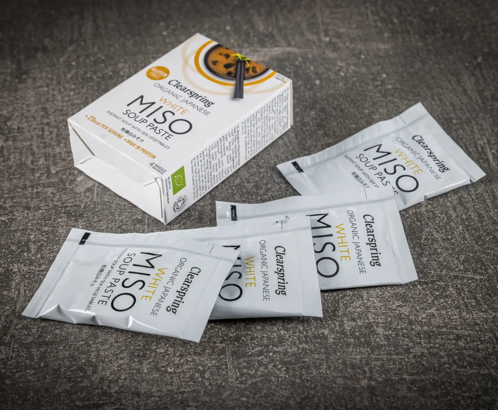 Eine weiße Schachtel Clearspring Bio Japanische Weiße Miso Suppenpaste mit vier Einzelportionen und mehrere lose Beutel derselben Instant-Suppe mit Meeresgemüse auf dunklem Untergrund. Die Packung ist glutenfrei und proteinreich.