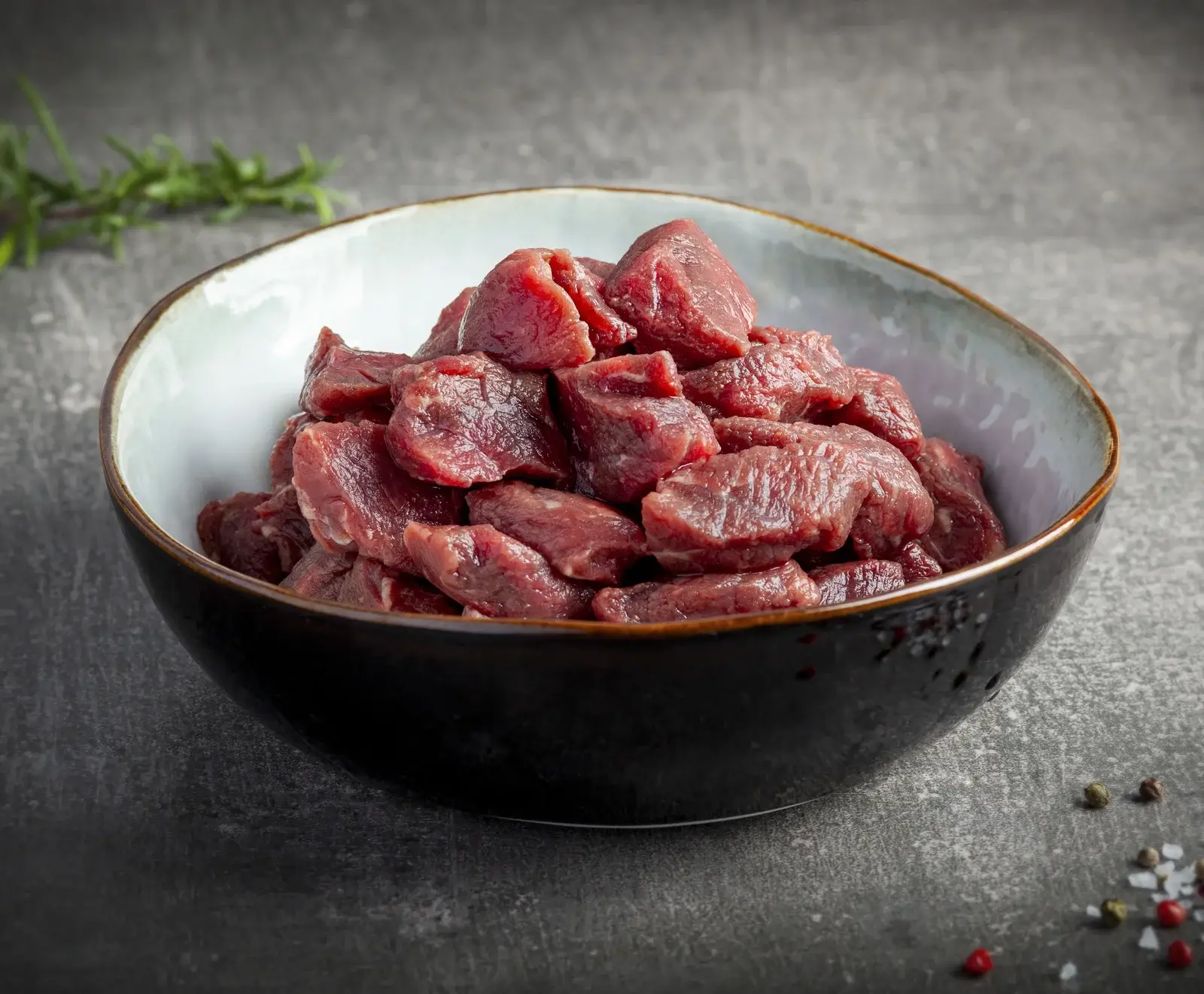Eine schwarze und hellgraue Schale gefüllt mit rohem, gewürfeltem Fleisch auf einem dunklen Untergrund. Im Hintergrund Rosmarin, im Vordergrund Pfefferkörner und Salz.