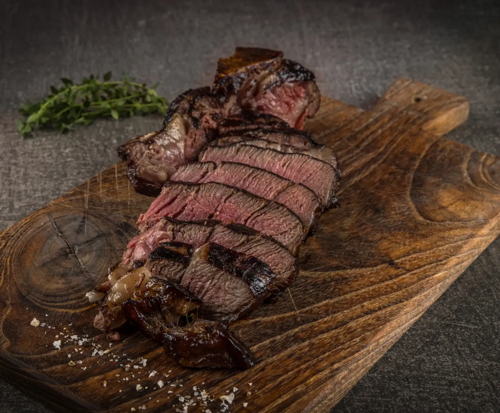 Ein aufgeschnittenes, medium-rare gegrilltes Steak auf einem rustikalen Holzbrett, bestreut mit grobem Salz und garniert mit Thymianzweigen auf dunklem Hintergrund.