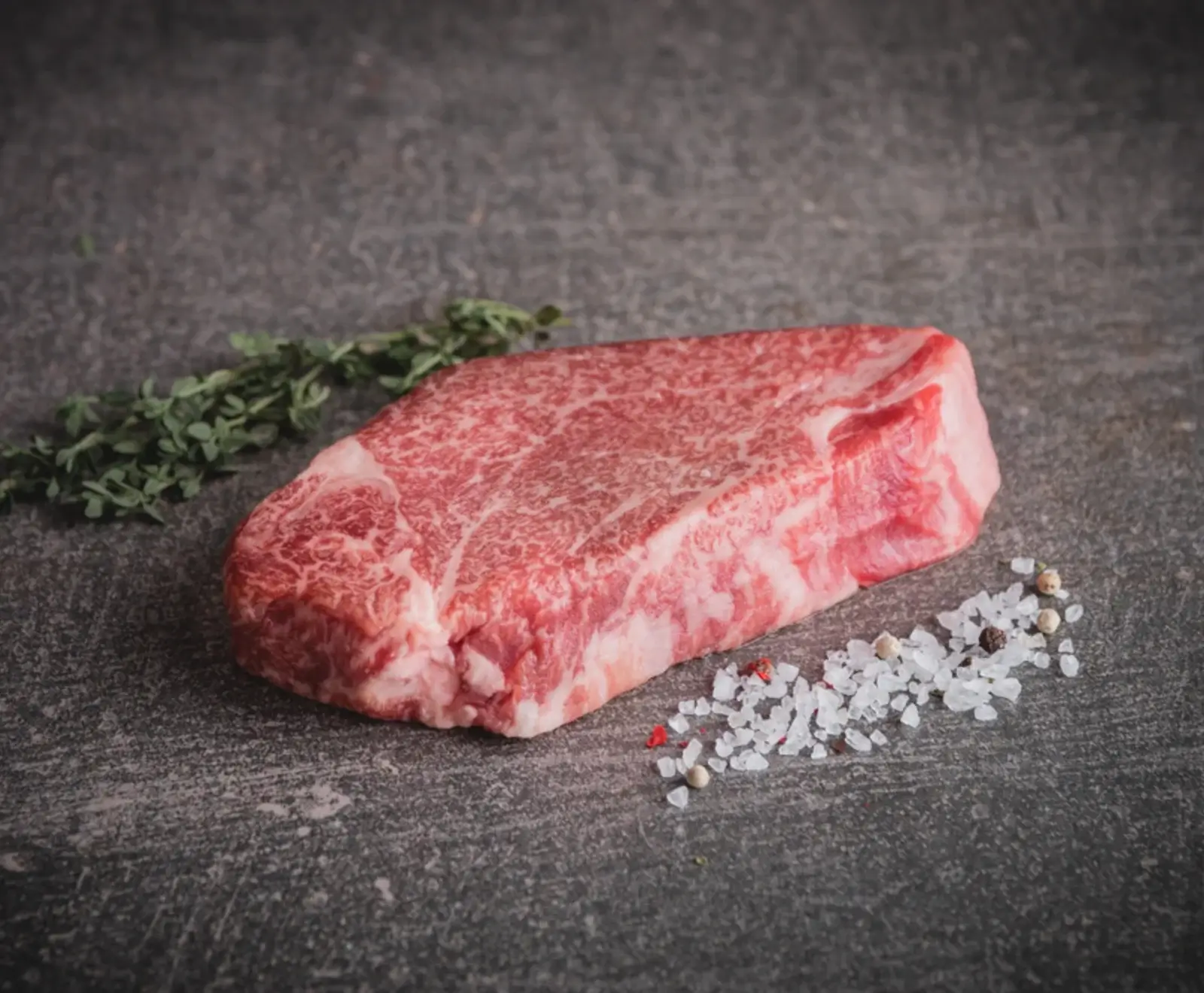 Ein rohes, stark marmoriertes Rindersteak liegt auf einer dunklen Oberfläche, daneben ein Zweig Thymian, grobes Meersalz und Pfefferkörner.