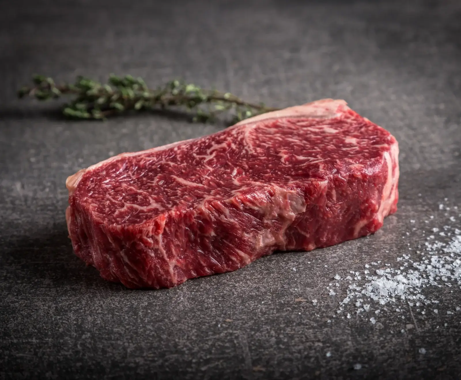 Ein rohes, stark marmoriertes Rindersteak liegt auf einer dunklen Steinplatte. Daneben liegen ein Thymianzweig und grobes Meersalz.