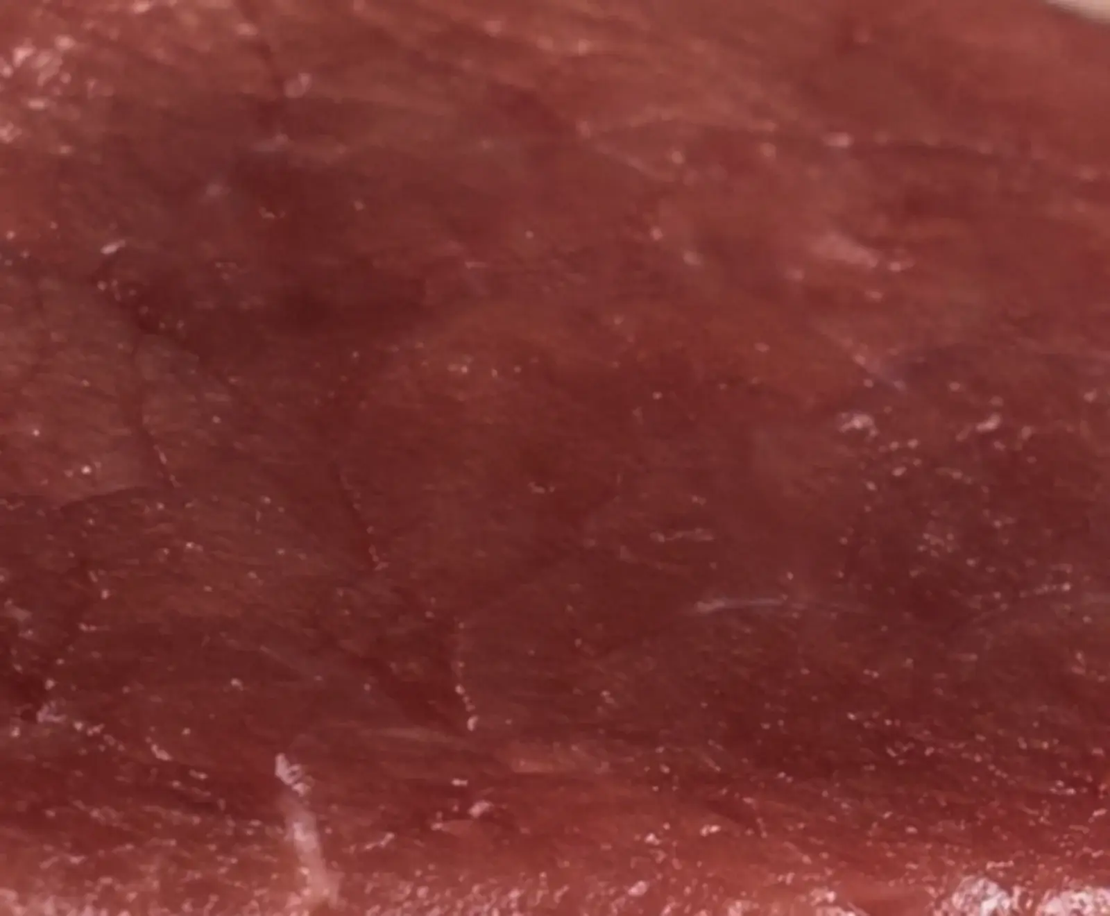 Nahaufnahme von tiefrotem, rohem Fleisch mit sichtbarer Faserstruktur und kleinen weißen Sprenkeln auf der leicht feuchten Oberfläche.