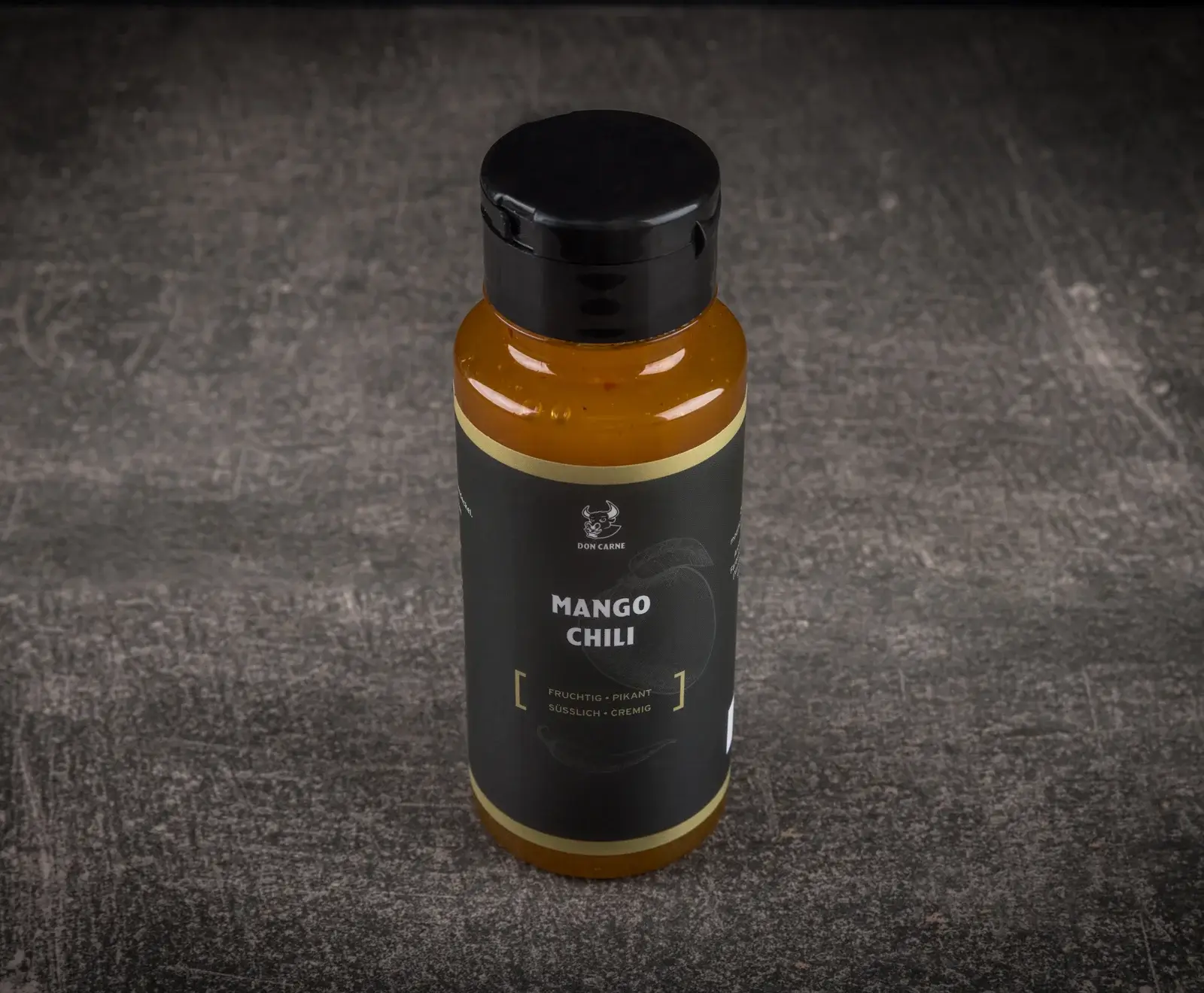 Eine Flasche Don Carne Mango Chili Sauce mit schwarzem Etikett und orangefarbenem Inhalt, stehend auf einem dunklen, strukturierten Untergrund. Das Etikett beschreibt die Sauce als fruchtig, pikant, süßlich und cremig.