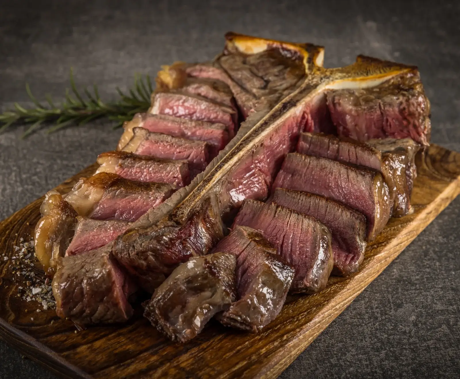 Ein perfekt gegartes, aufgeschnittenes T-Bone-Steak, medium rare, liegt auf einem rustikalen Holzbrett. Die rosa Fleischscheiben sind mit grobem Salz bestreut, ein Rosmarinzweig liegt im Hintergrund.