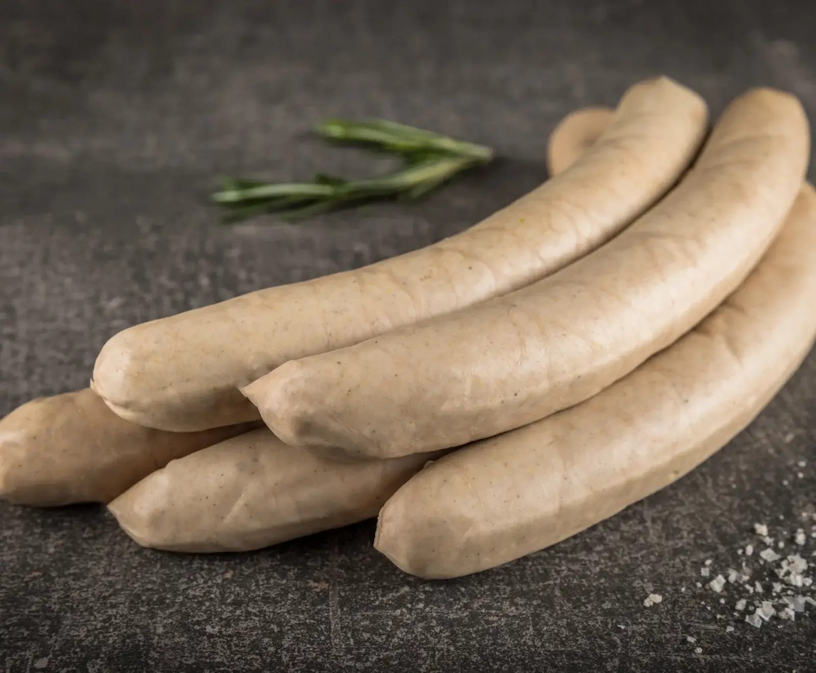 Mehrere rohe, helle Würste liegen auf einem dunklen, strukturierten Untergrund. Im unscharfen Hintergrund sind grüne Rosmarinzweige zu sehen, im Vordergrund grobe Salzkristalle.