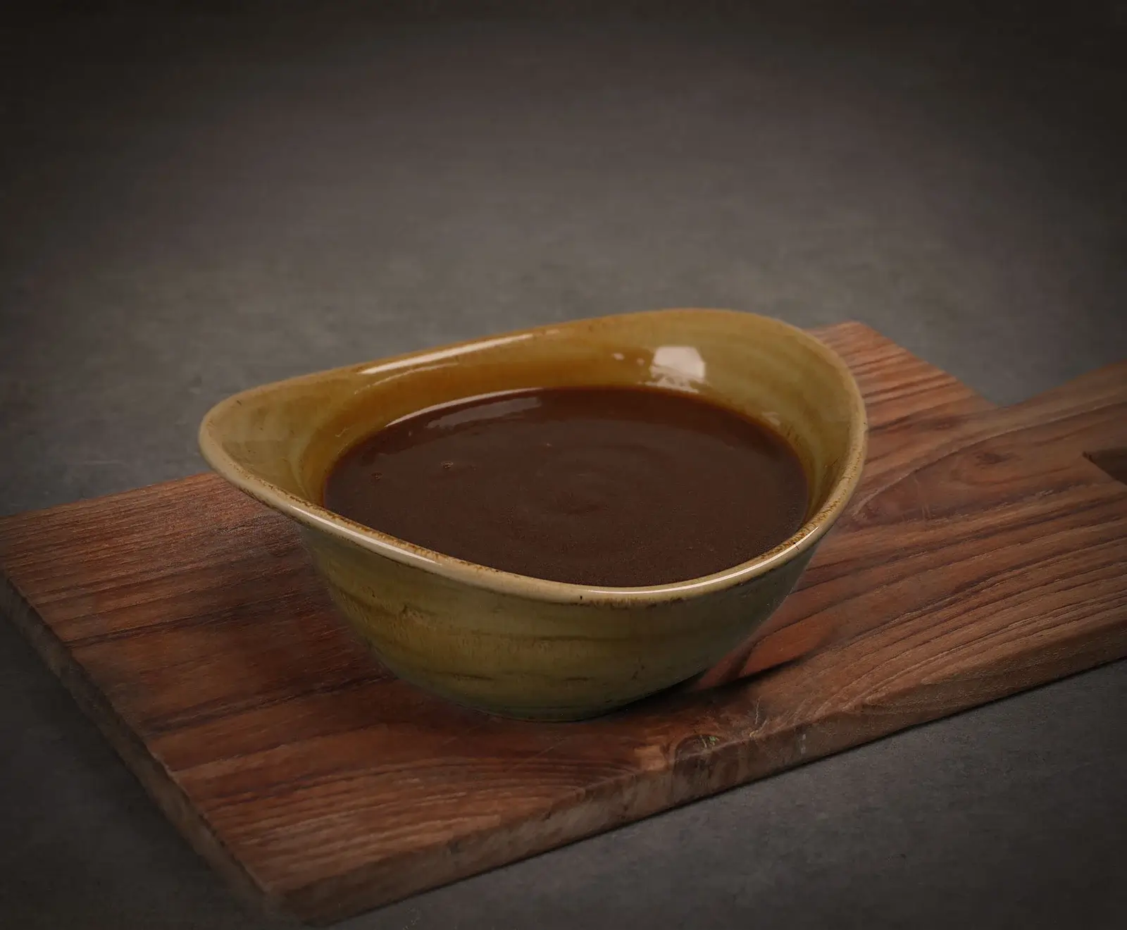 Dunkelbraune Soße in einer beigen Keramikschale mit unregelmäßigem Rand, platziert auf einem Holzschneidebrett vor dunklem Hintergrund.