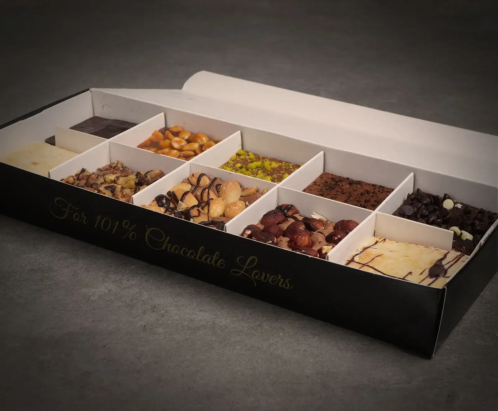 Eine offene schwarze Geschenkbox mit weißen Unterteilungen, gefüllt mit zehn verschiedenen Schokoladen- und Nussspezialitäten, darunter Macadamias, Haselnüsse, Pistazien und Schokostückchen. Die Box trägt die Aufschrift "For 101% Chocolate Lovers".