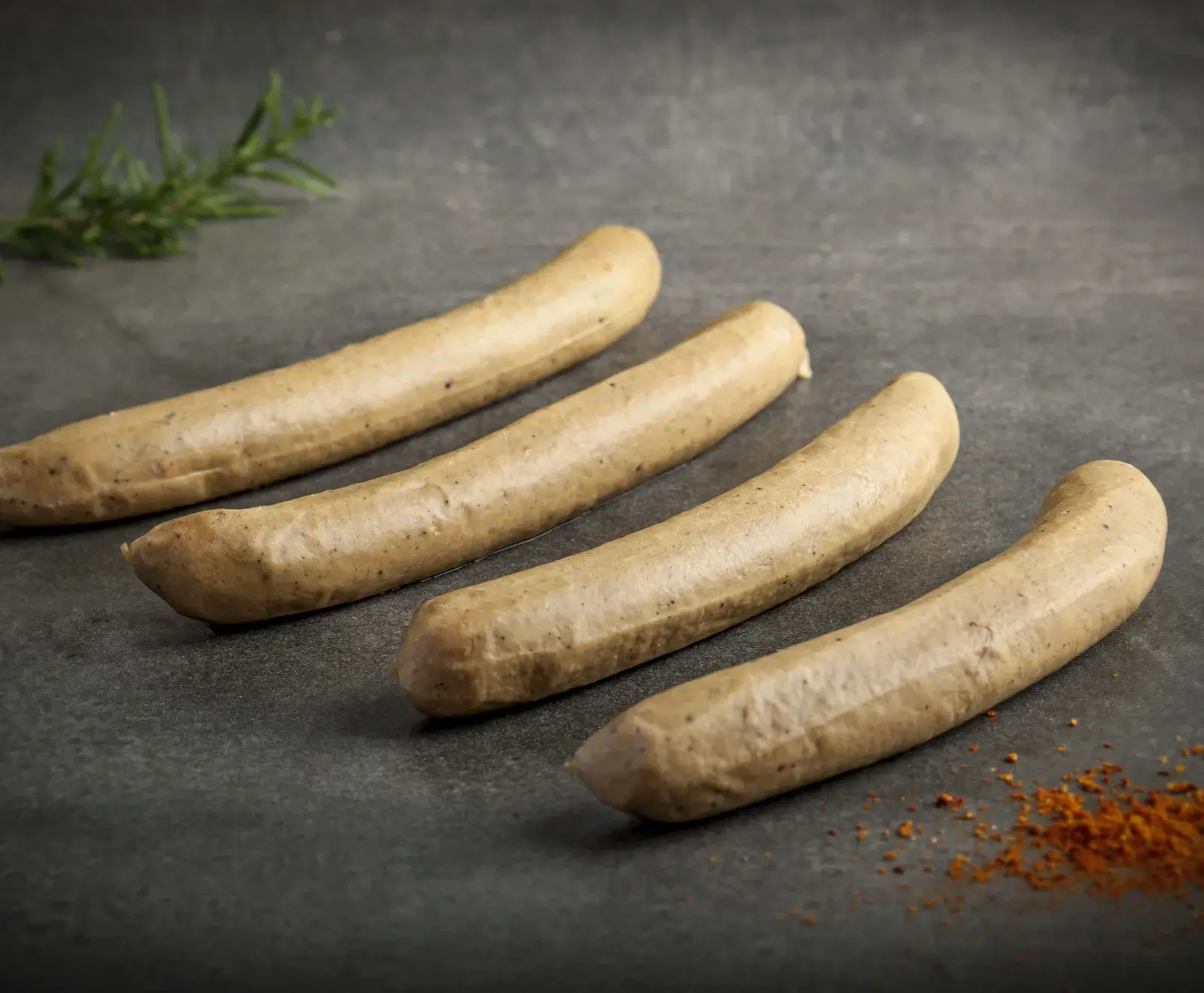 Vier ungekochte, hellbraune Würste mit dunklen Sprenkeln liegen diagonal auf einer dunklen, strukturierten Oberfläche. Ein Rosmarinzweig und Paprikapulver sind im Bild.