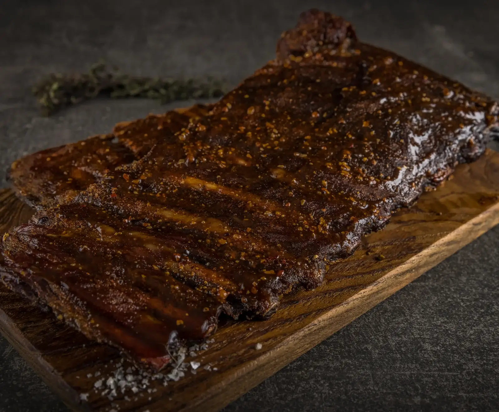 Ein großes, glasiertes Rippchenstück auf einem Holzbrett, bedeckt mit dunkler, glänzender BBQ-Sauce und sichtbaren Gewürzen. Grobes Salz liegt am unteren Rand, im Hintergrund unscharfe Kräuter.