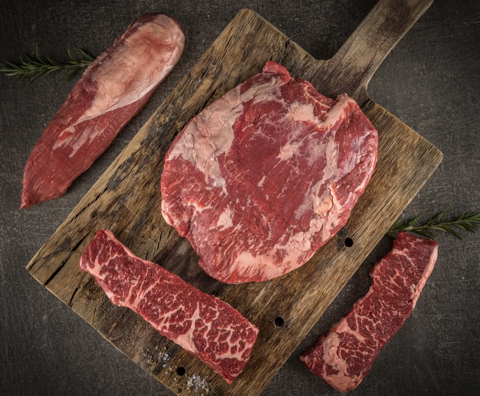 Verschiedene Schnitte von rohem Rindfleisch, einschließlich Filet, Ribeye und Flankensteak, auf einem rustikalen Holzbrett mit Rosmarin.