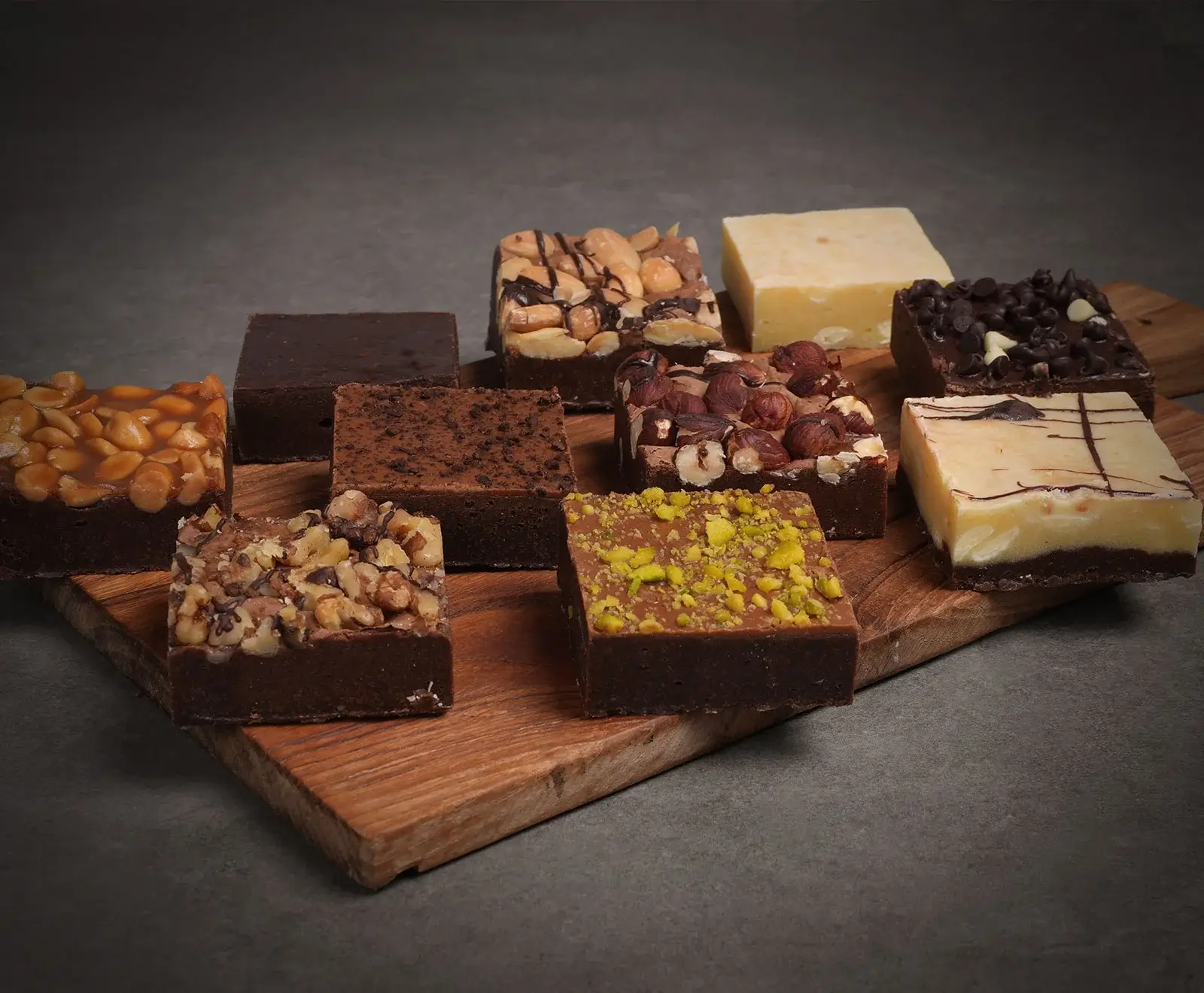 Zehn quadratische Brownies und Fudge-Stücke mit vielfältigen Belägen wie Erdnüssen, Haselnüssen, Walnüssen, Pistazien, Schokochips und Karamell, appetitlich auf einem Holzbrett arrangiert.