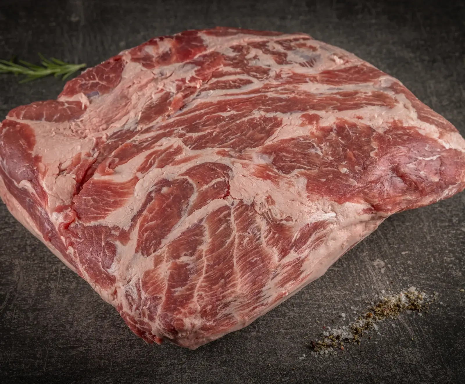 Ein rohes Stück marmoriertes Rindfleisch auf einer dunklen Oberfläche, mit Rosmarin als Garnitur