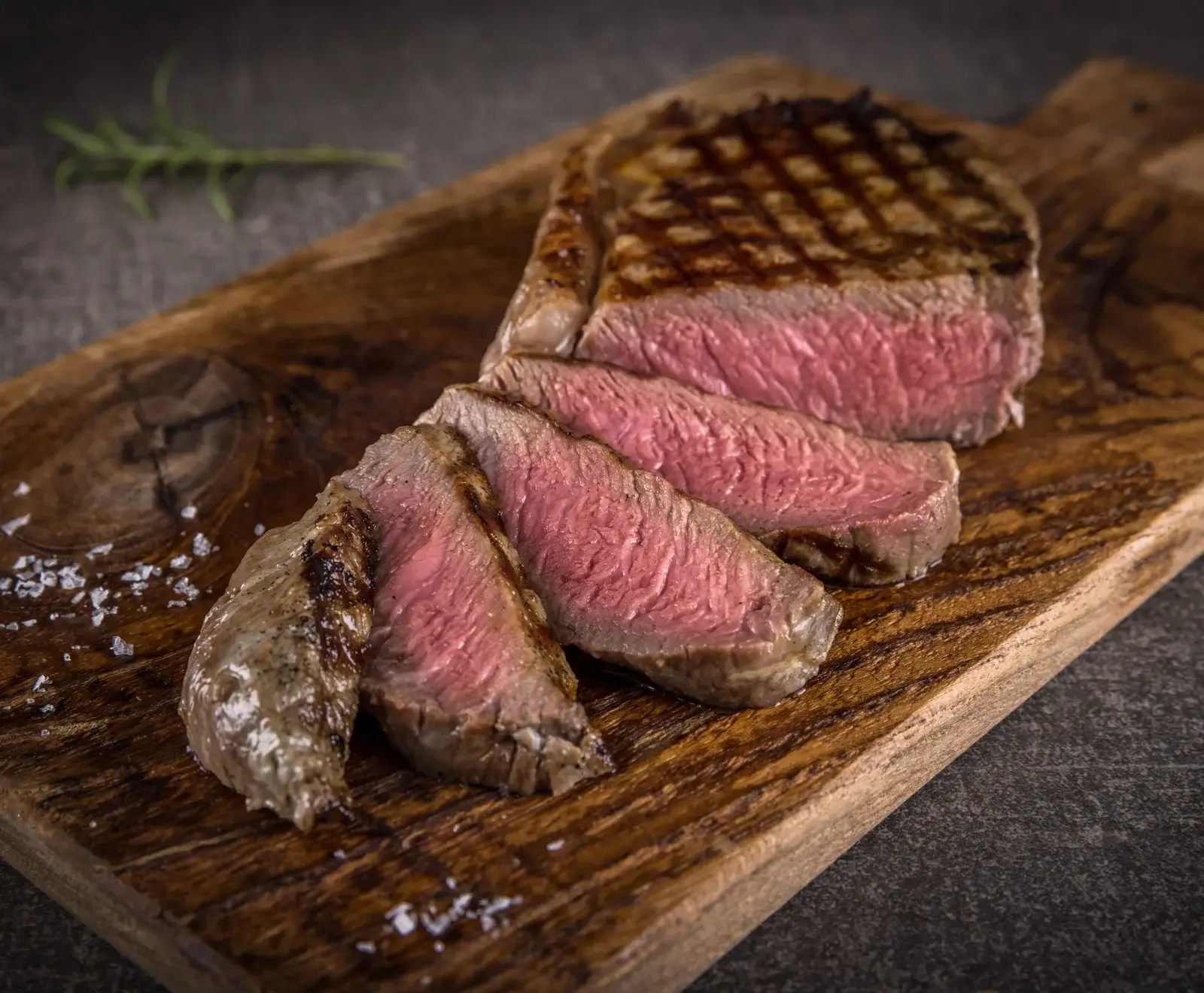 Nahaufnahme eines saftigen, medium-rare gegrillten Steaks, in Scheiben geschnitten, auf einem rustikalen Holzbrett mit grobem Meersalz und Rosmarin.