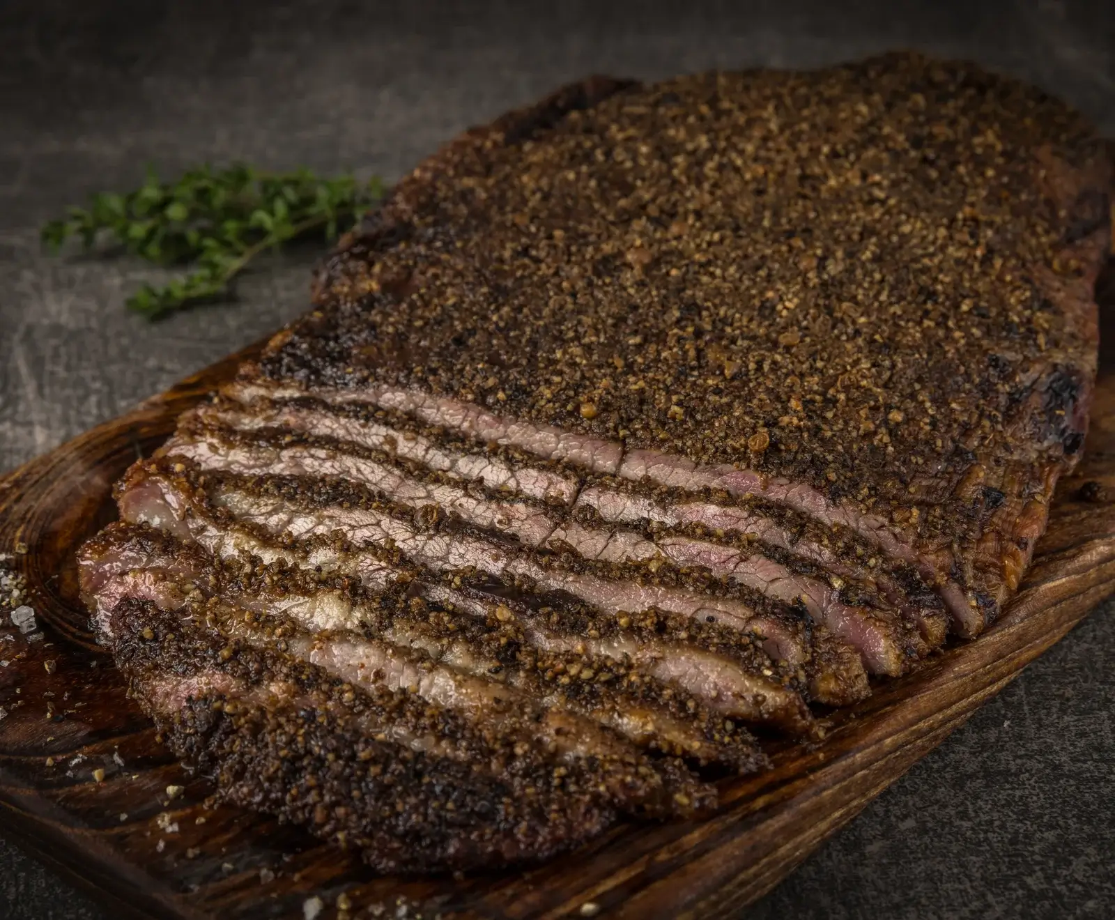 Ein großes, geschnittenes Stück Brisket mit einer dicken, dunklen Pfefferkruste liegt auf einem Holzbrett. Die Scheiben zeigen das zarte, rosa Fleisch. Im Hintergrund sind Kräuter und grobes Salz zu sehen.