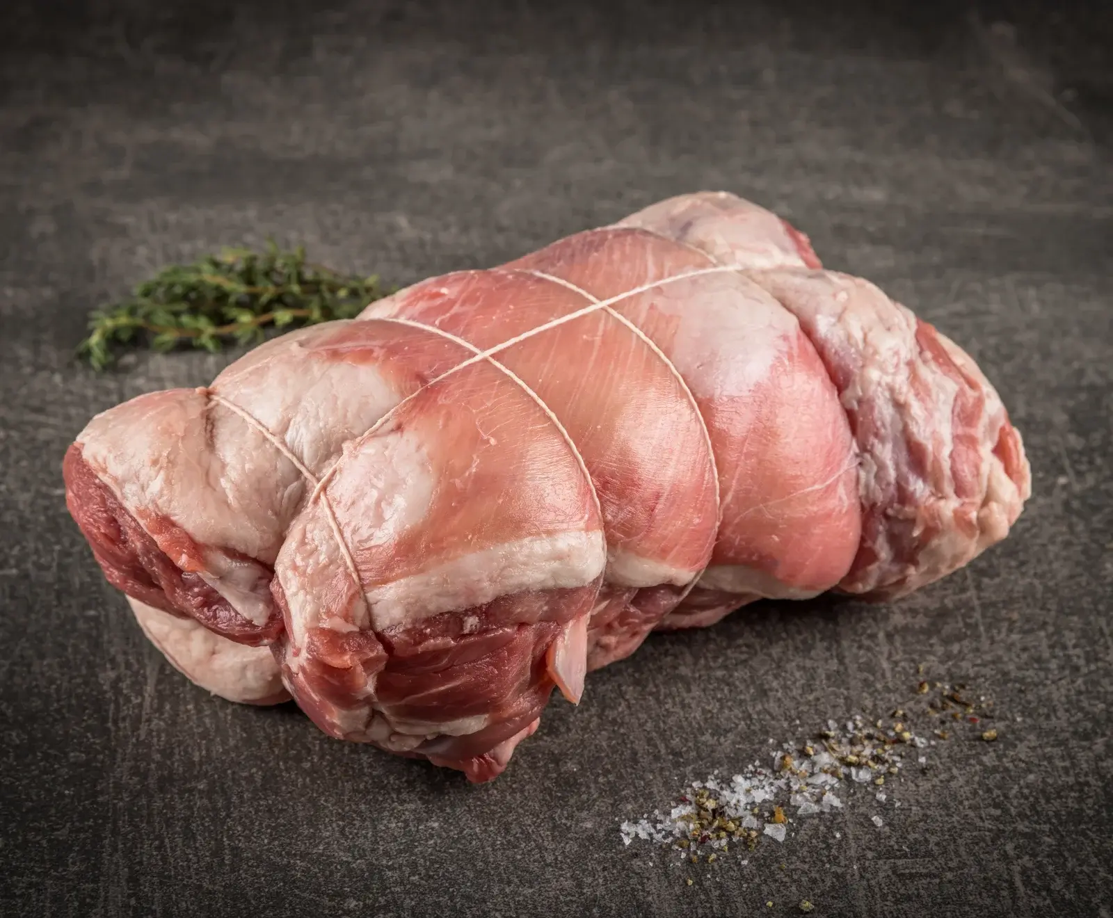 Ein roher, mit weißem Küchengarn gebundener Braten, mit sichtbarem Fett und magerem Fleisch, liegt auf einer dunklen Oberfläche. Daneben sind frischer Thymian, grobes Salz und Pfeffer.