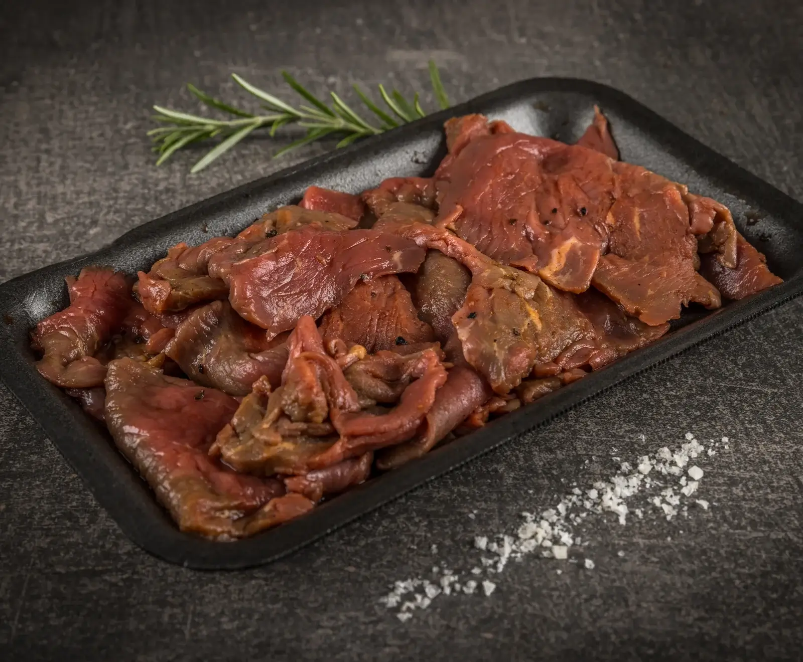 Rohe, marinierte Fleischstreifen in einer schwarzen Schale, daneben ein Rosmarinzweig und grobes Salz auf dunklem Untergrund.