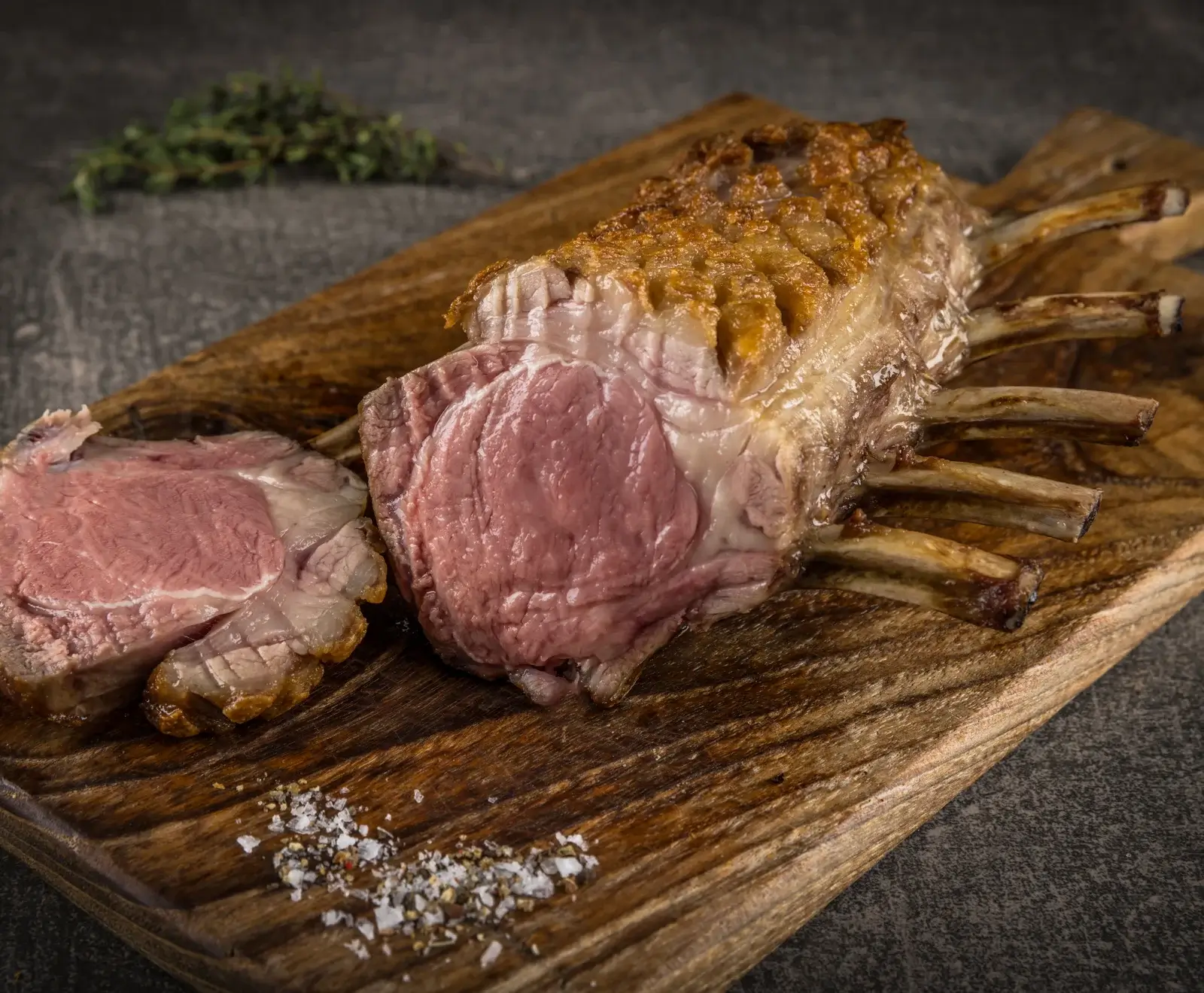 Ein saftiges, medium-rare gebratenes Lammkarree auf einem Holzschneidebrett, mit einer Scheibe daneben, bestreut mit grobem Salz und Pfeffer. Im Hintergrund Thymian.