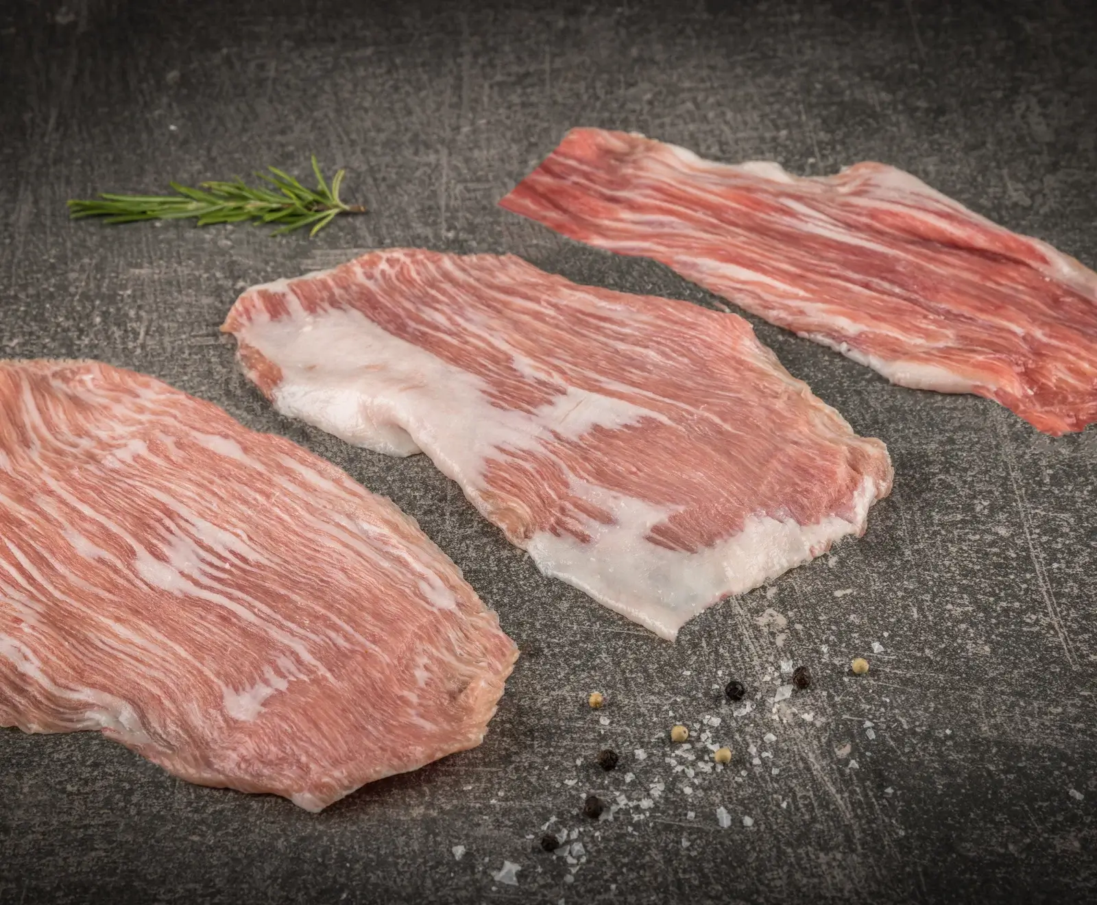 Rohe, dünn geschnittene Fleischstücke mit Fettmarmorierung auf dunklem Stein, dazu Rosmarin, Pfeffer und Salz.