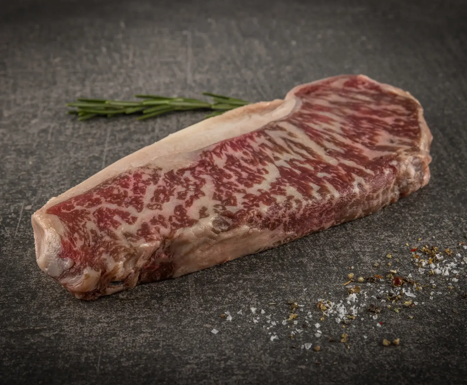 Ein rohes, stark marmoriertes Wagyu-Rindersteak (Strip Loin) liegt auf einer dunklen Steinplatte, daneben ein Rosmarinzweig, grobes Salz und Pfeffer.