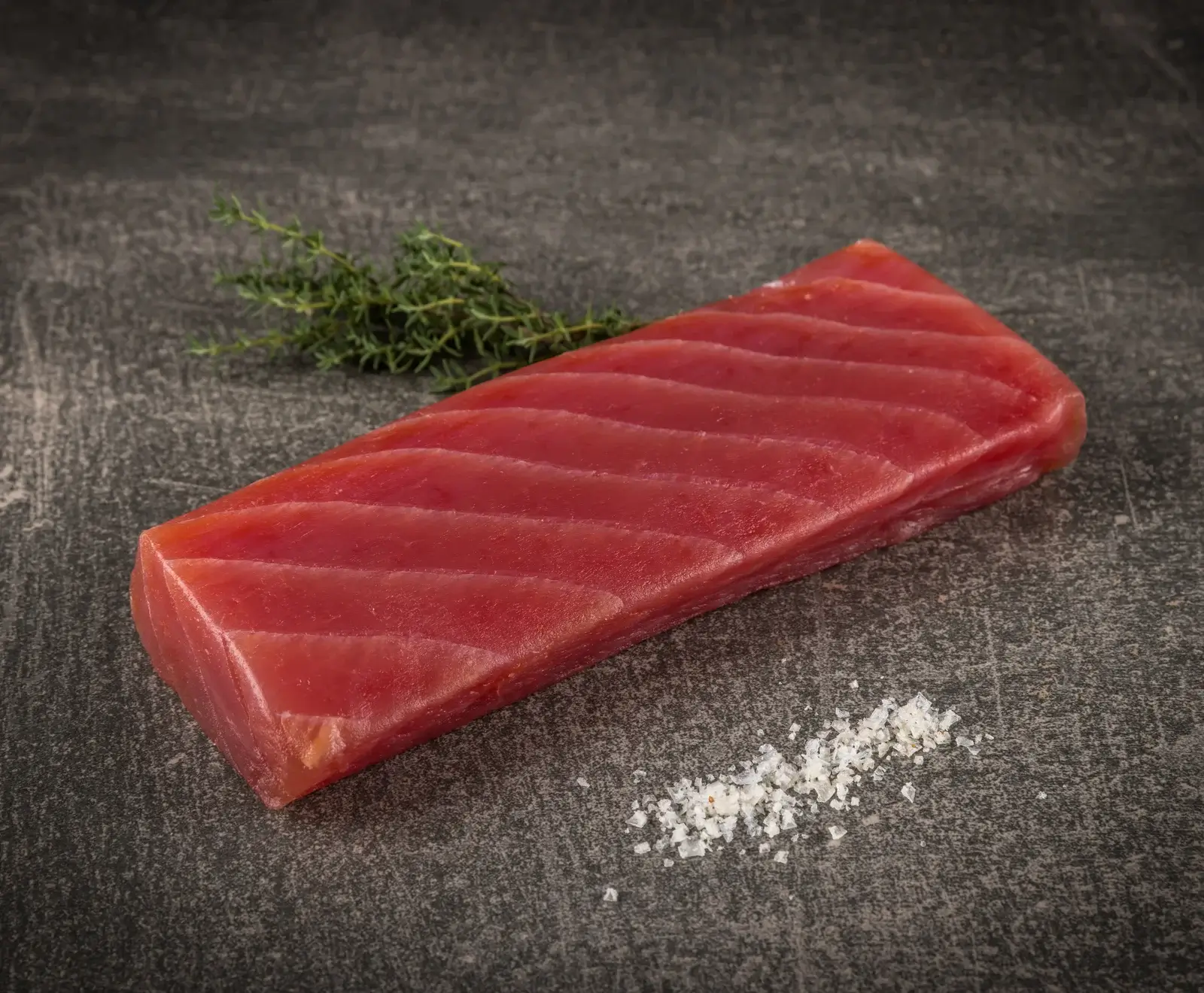 Ein rohes, leuchtend rotes Thunfischfiletstück liegt auf einer dunklen Steinoberfläche, begleitet von einem Zweig frischem Thymian und grobem Meersalz.