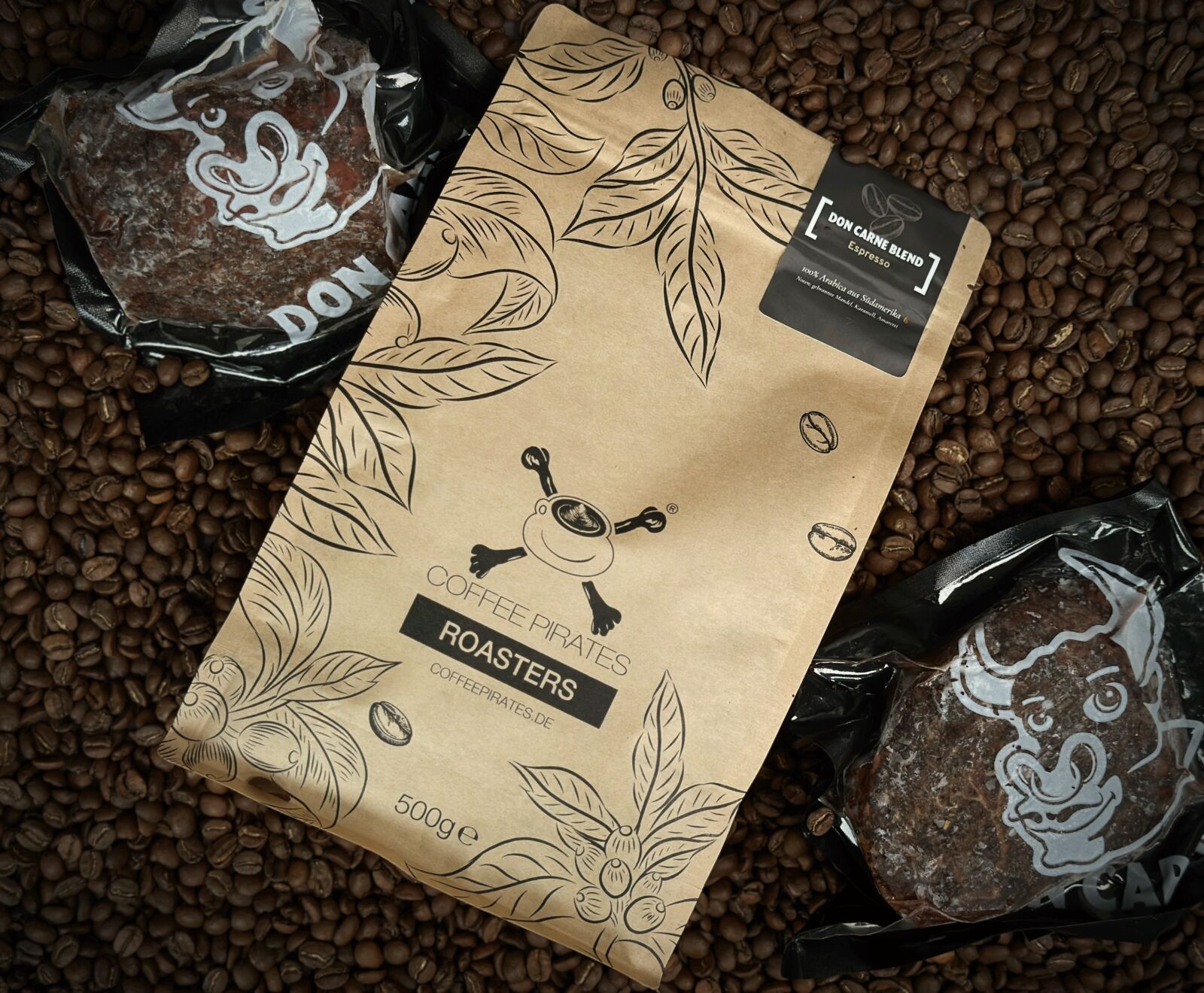 Eine braune Kaffeepackung von Coffee Pirates Roasters (Don Carne Blend Espresso, 500g) liegt auf Kaffeebohnen, flankiert von zwei vakuumverpackten Don Carne Fleischstücken in Stierkopfform.