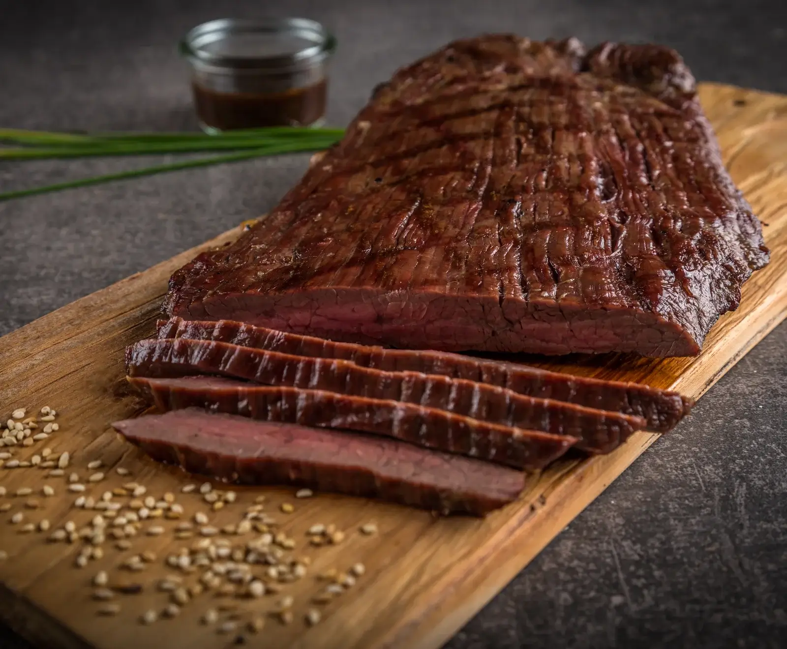 Ein aufgeschnittenes, gegrilltes Flanksteak mit rosa Kern auf einem Holzbrett, umgeben von Sesamkörnern, mit einer Soße und Schnittlauch im unscharfen Hintergrund.