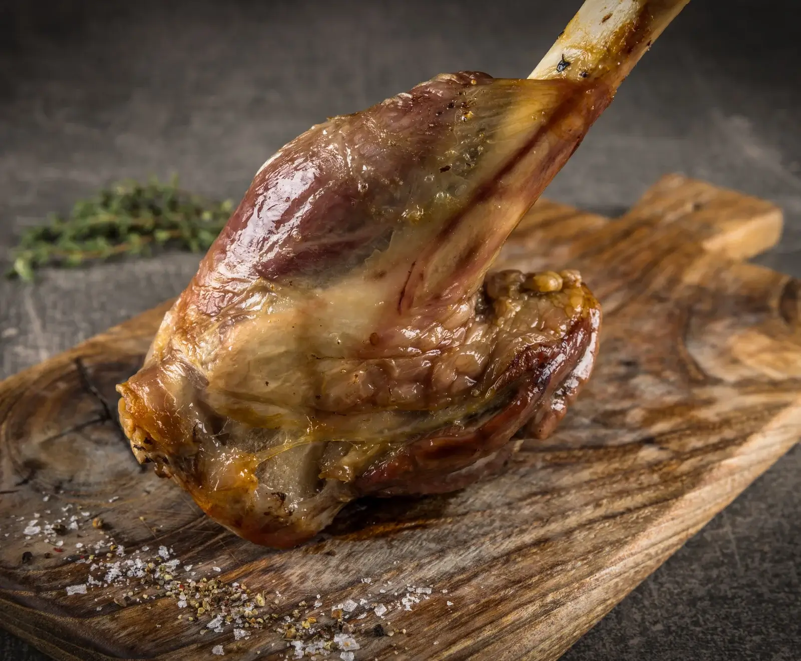 Eine goldbraun gebratene Lammkeule liegt auf einem rustikalen Holzbrett, bestreut mit Salz und Pfeffer. Im unscharfen Hintergrund ist frischer Thymian zu sehen.