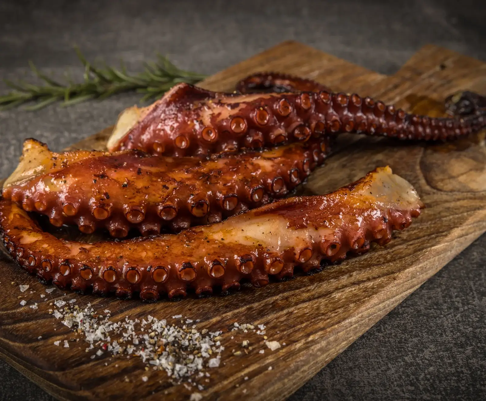 Gegrillte Oktopusarme auf einem rustikalen Holzbrett, glänzend rötlich-braun mit sichtbaren Saugnäpfen. Grobes Salz, Pfeffer und ein Rosmarinzweig sind im Bild zu sehen.
