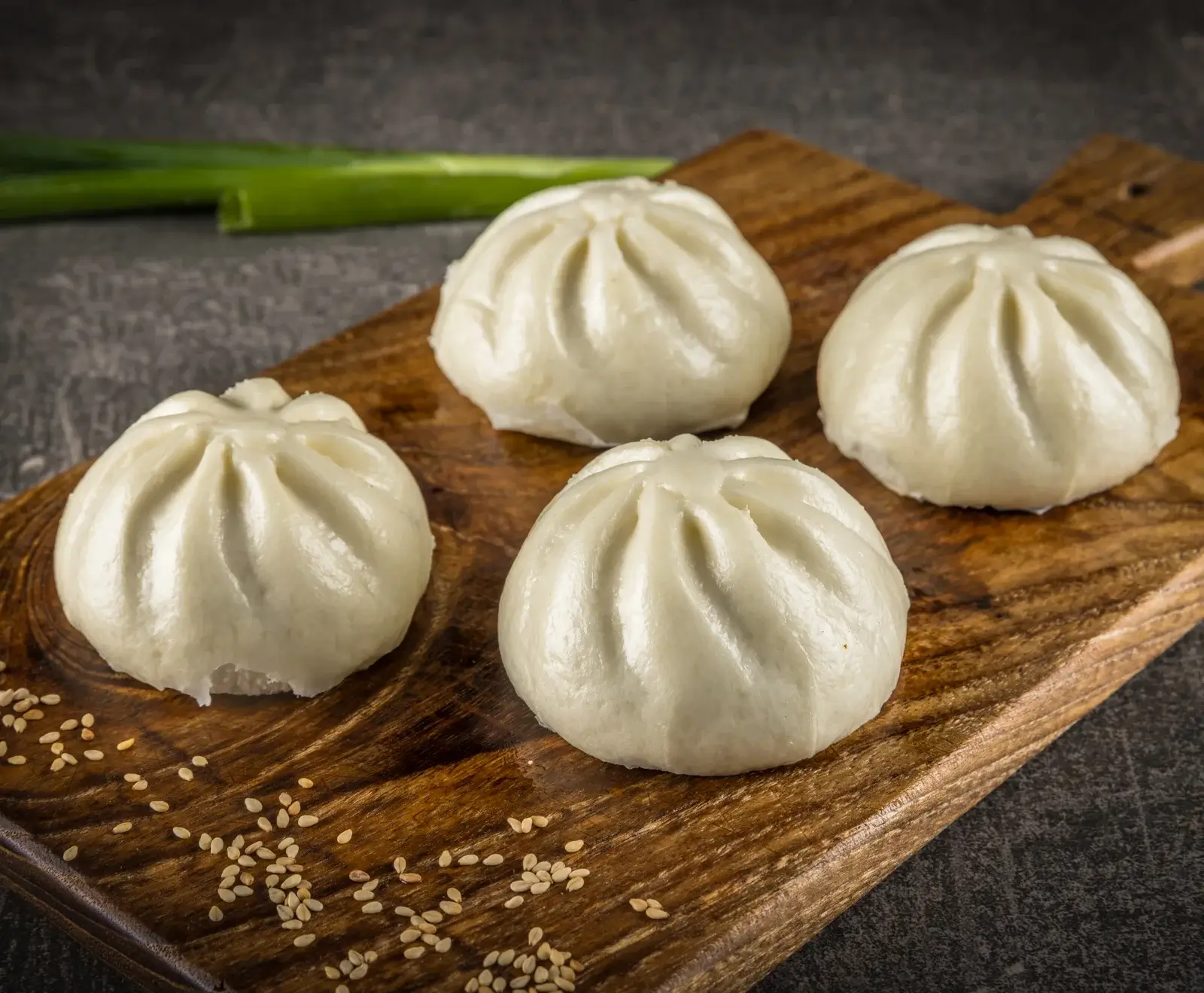 Vier weiße, gedämpfte Baozi-Brötchen mit gefalteten Oberseiten liegen auf einem rustikalen Holzbrett. Sesamsamen sind verstreut, grüne Zwiebeln im dunklen Hintergrund.