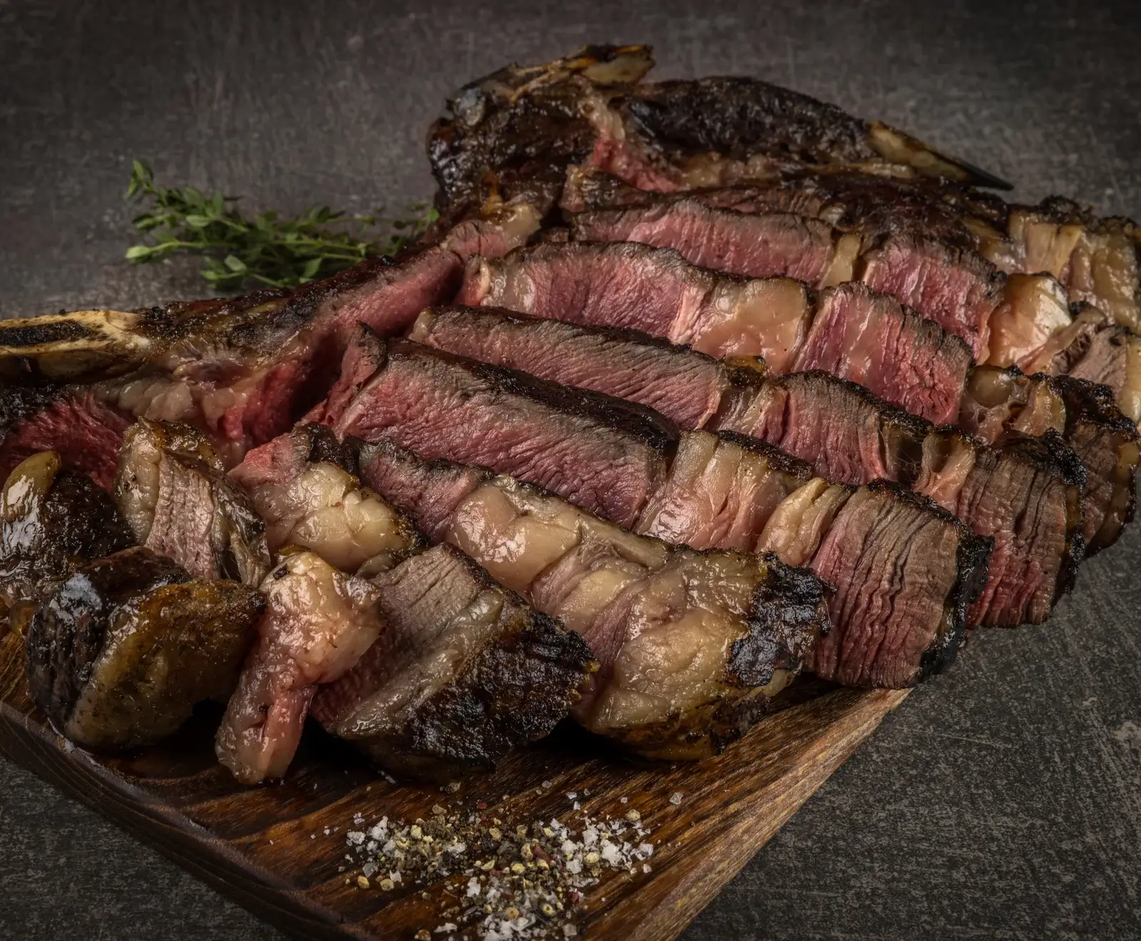 Ein perfekt medium-rare gegartes, in Scheiben geschnittenes Tomahawk-Steak auf einem Holzbrett, garniert mit grobem Salz, Pfeffer und einem Thymianzweig.